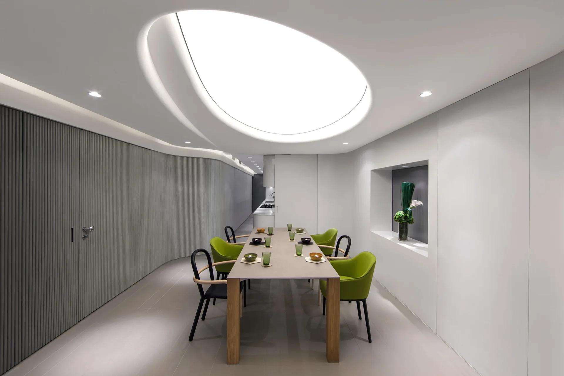 A minimalist dining room featuring a long dining table with green chairs set beneath a large illuminated oval ceiling light and next to a curved fluted wall.