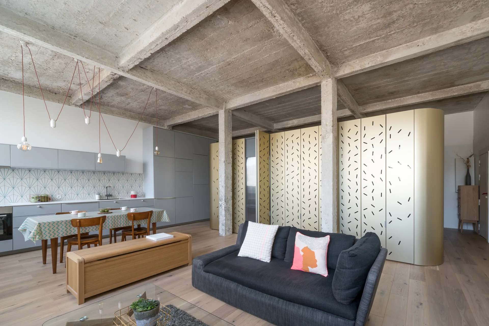 An open-plan living room and kitchen featuring an exposed concrete ceiling, a dark grey sofa, and tall gold-patterned decorative screens.