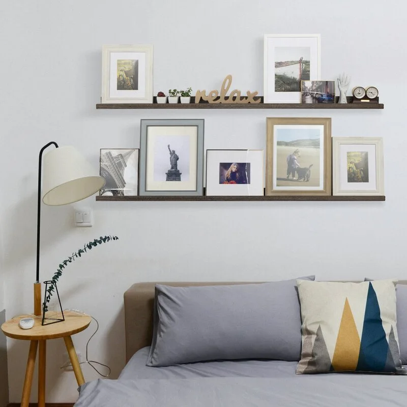 Two wooden picture ledges holding framed photos hang on a white wall above a bed with grey sheets and a round wooden side table.