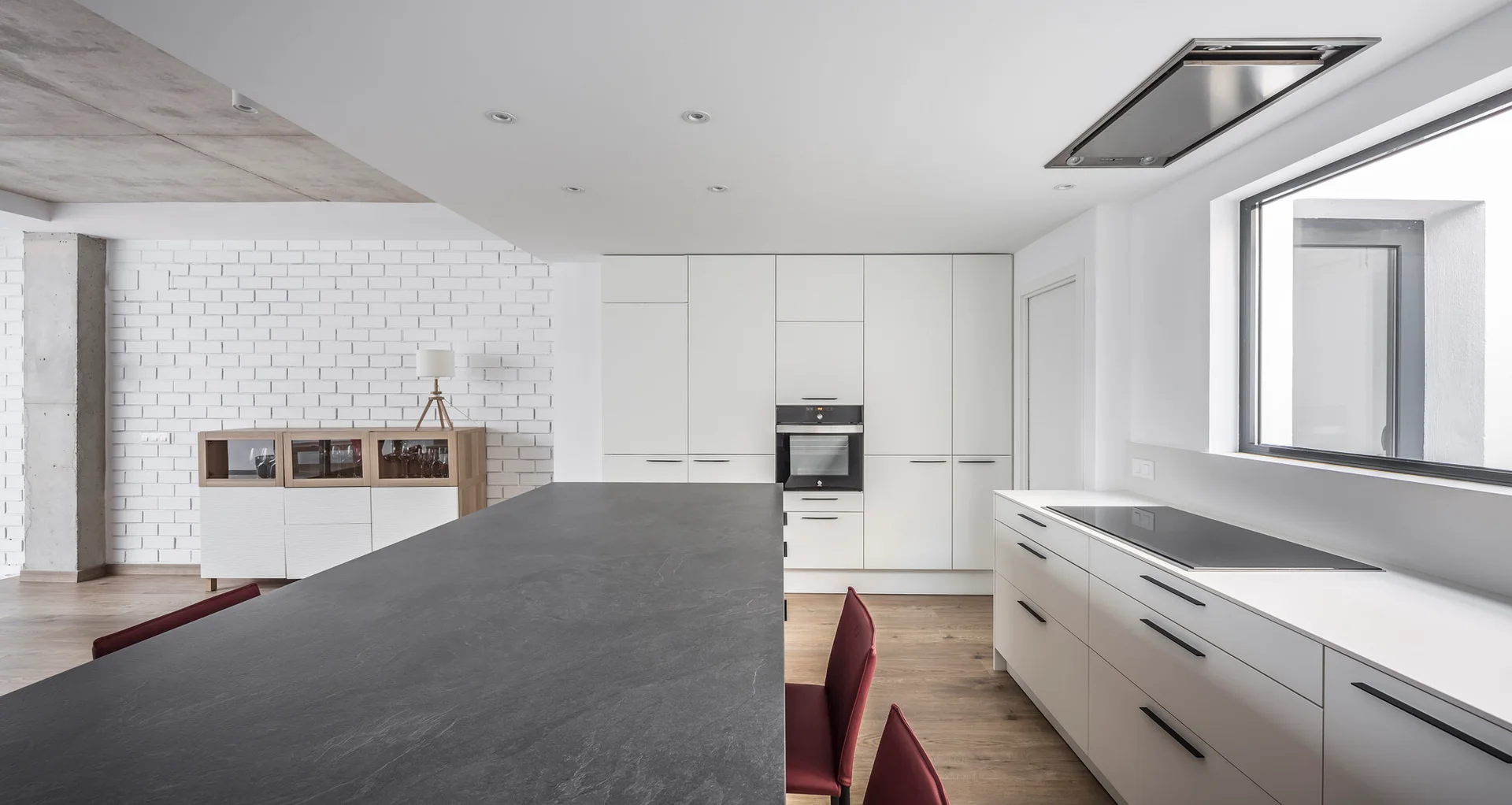 A modern minimalist kitchen featuring a long charcoal stone island with red chairs, flat-front white cabinets, and a painted white brick wall.
