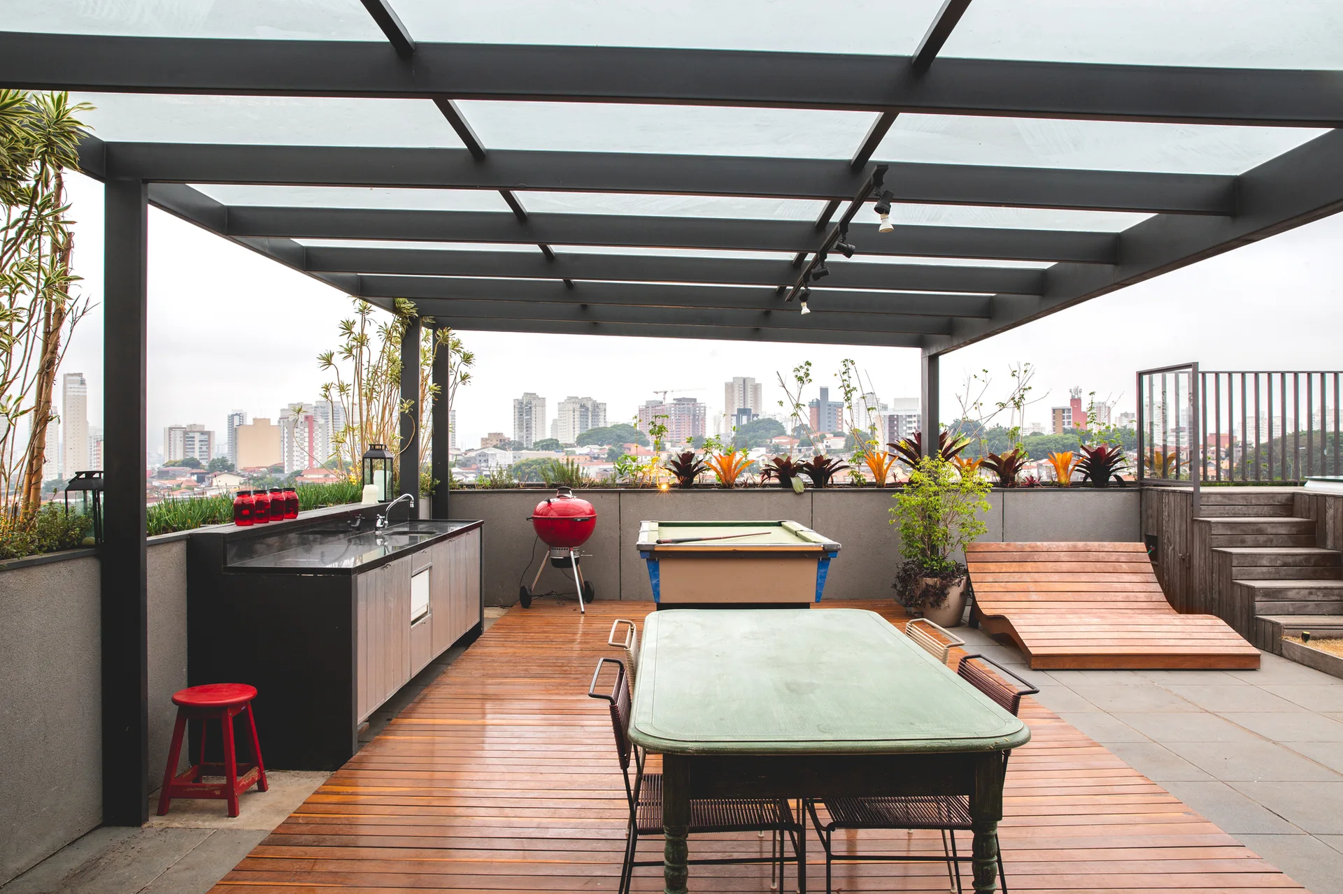 A rooftop terrace featuring a wood plank deck under a black metal pergola, an outdoor kitchen, a green dining table, and a pool table.