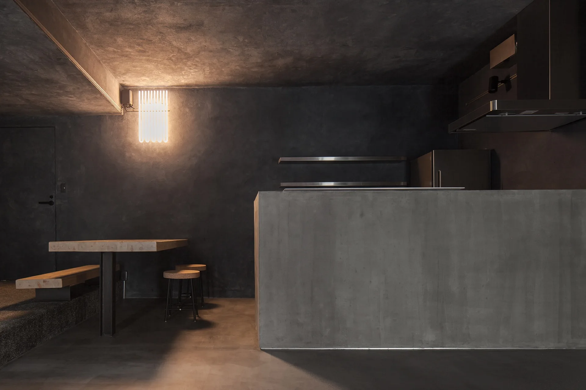 A dark minimalist kitchen features a seamless concrete island, dark grey textured walls, and a raw wood dining table with matching bench.