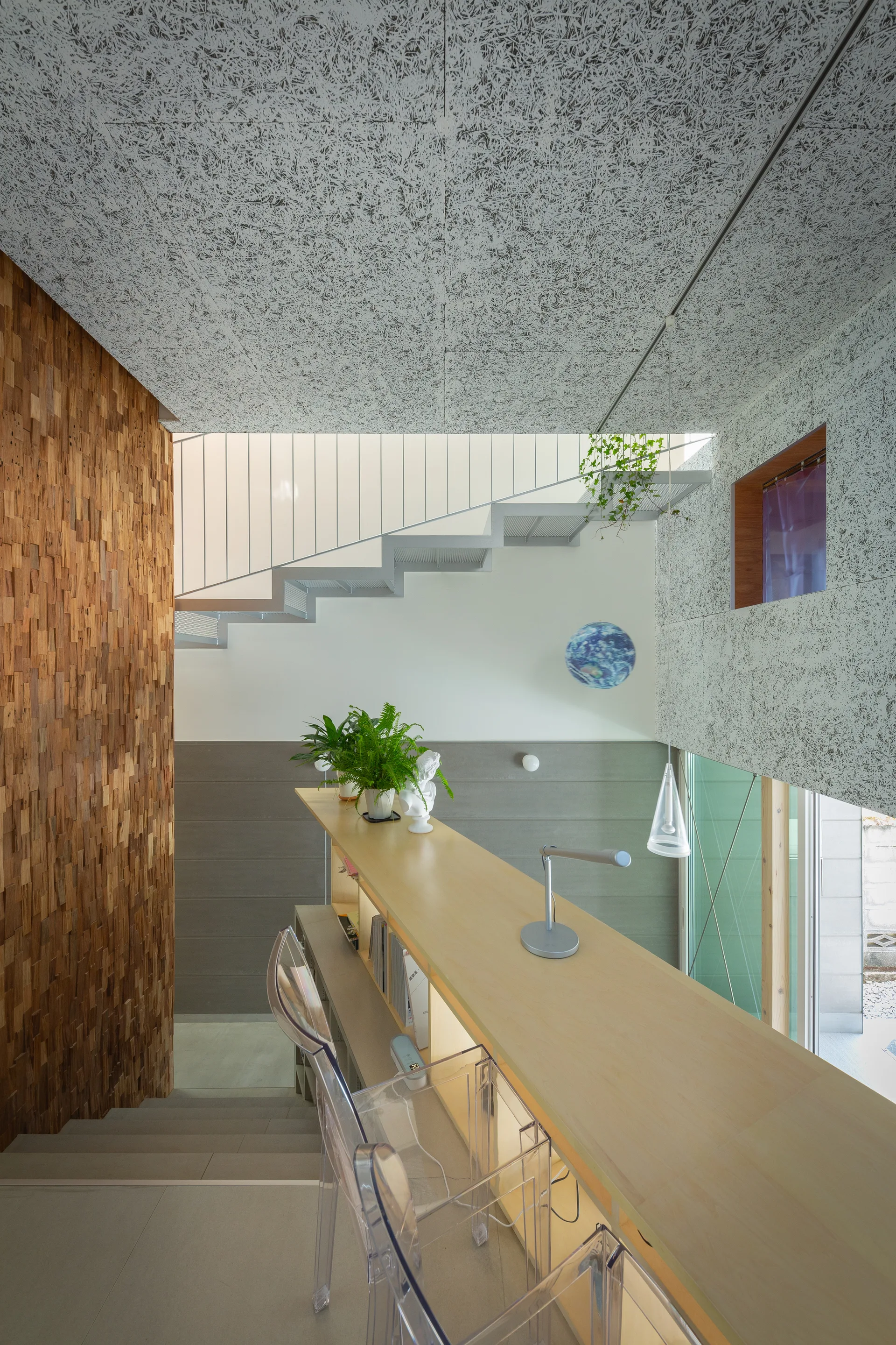 A multi-level interior workspace featuring a long light wood counter with clear acrylic stools, an open metal staircase, and a textured wood-block wall.