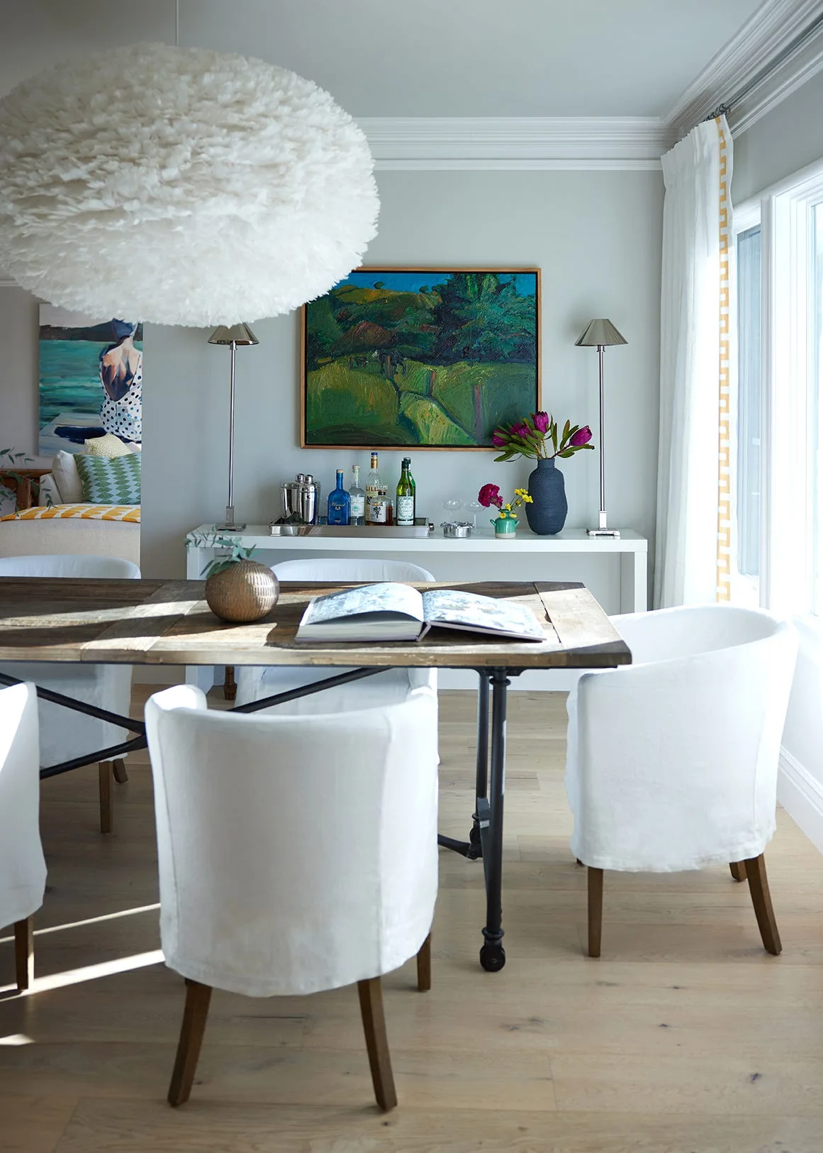 White slipcovered dining chairs surround a rustic wooden table beneath a fluffy feathered pendant light in a bright dining room.