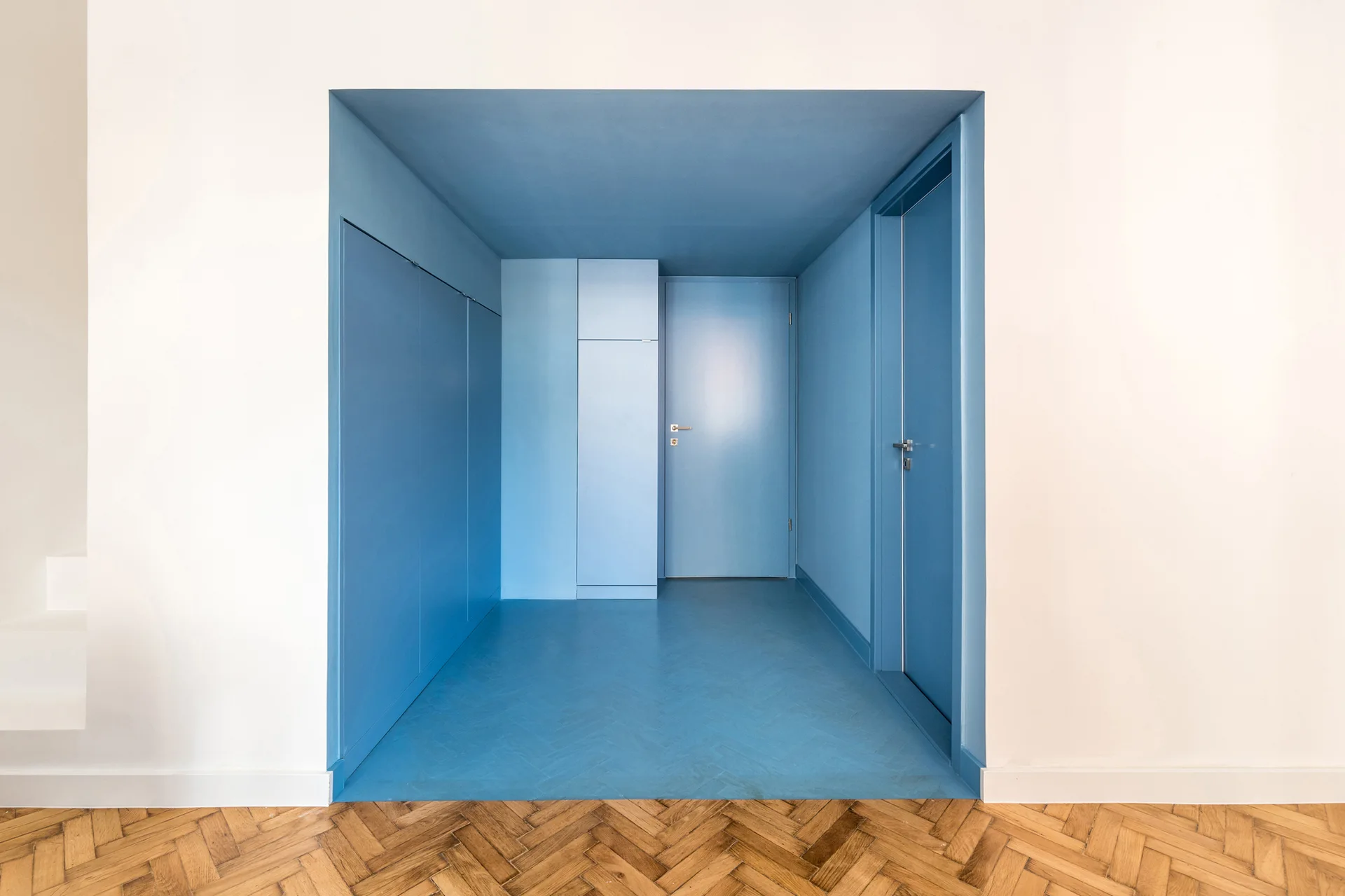 A recessed hallway completely painted in matte blue features integrated storage and matching doors, contrasting with warm herringbone parquet flooring.