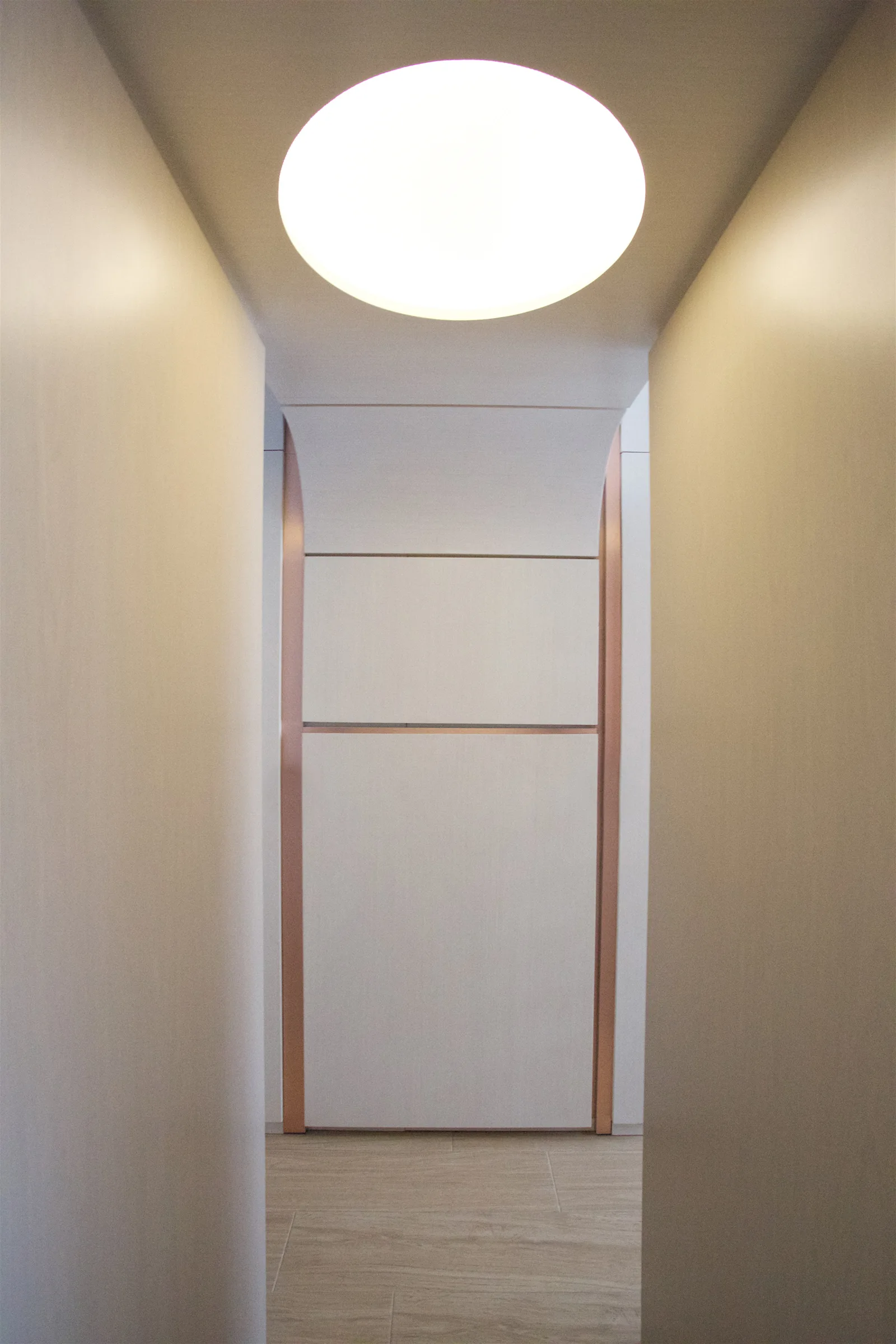A narrow modern hallway with light wood flooring features a large circular ceiling light and a paneled accent wall with metallic trim.