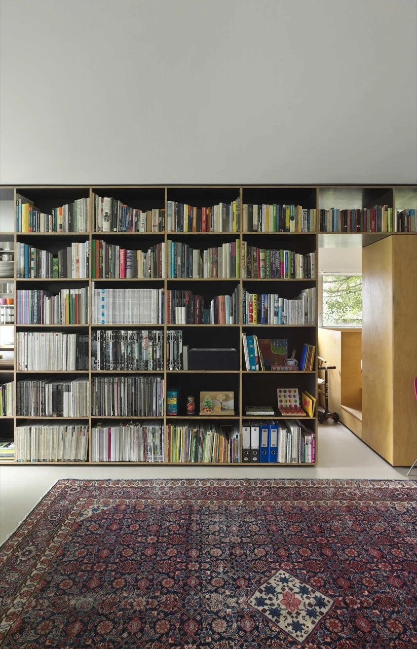 A large floor-to-ceiling dark bookshelf filled with books stands next to a wooden doorway, with an intricate red and blue patterned rug on the white floor.