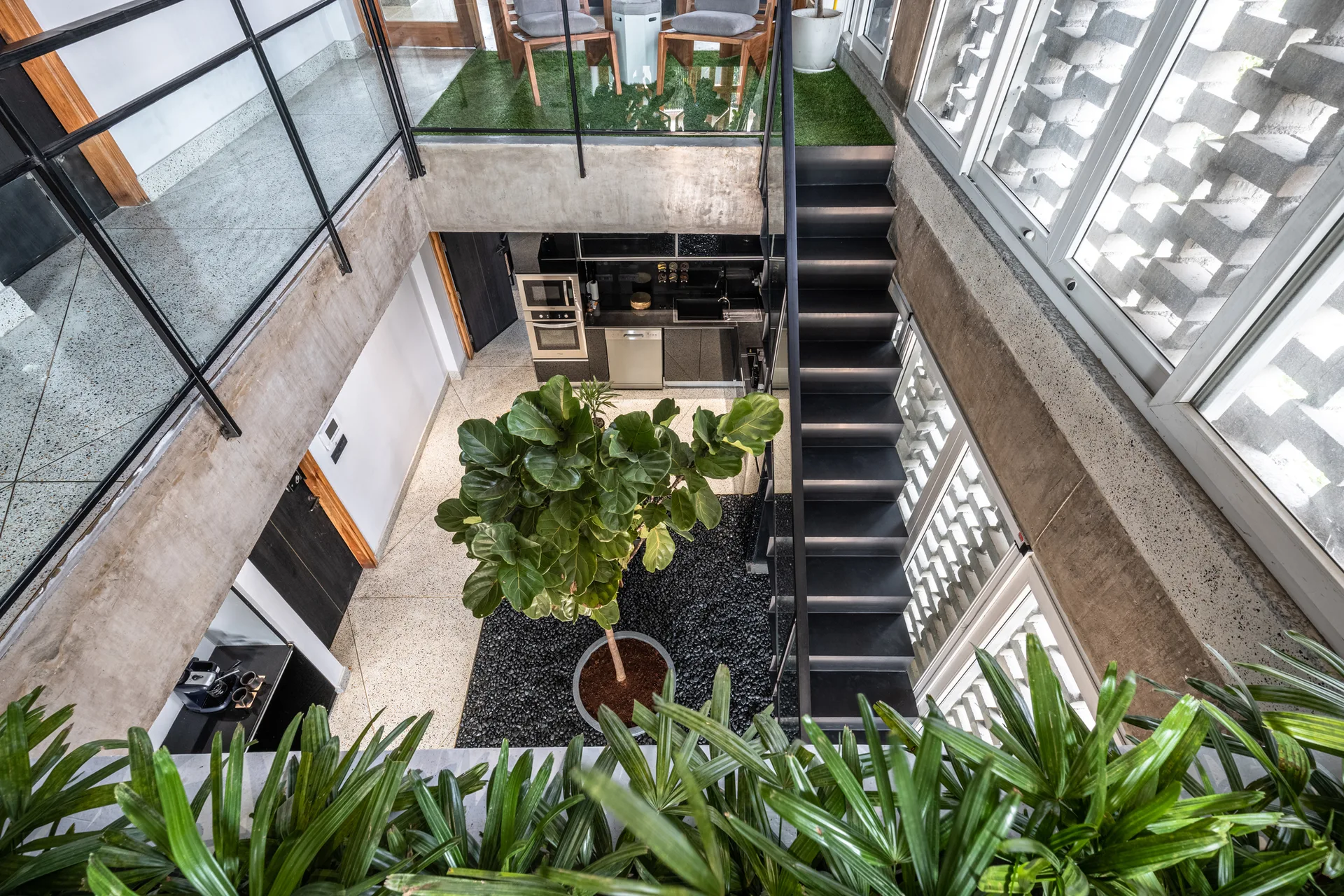 A high-angle view of a multi-level concrete atrium featuring a central green tree in a black gravel bed next to a black metal staircase.