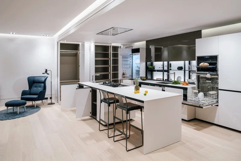 A modern white kitchen features a central island, integrated appliances, and a blue armchair with a matching ottoman on a light wood floor.