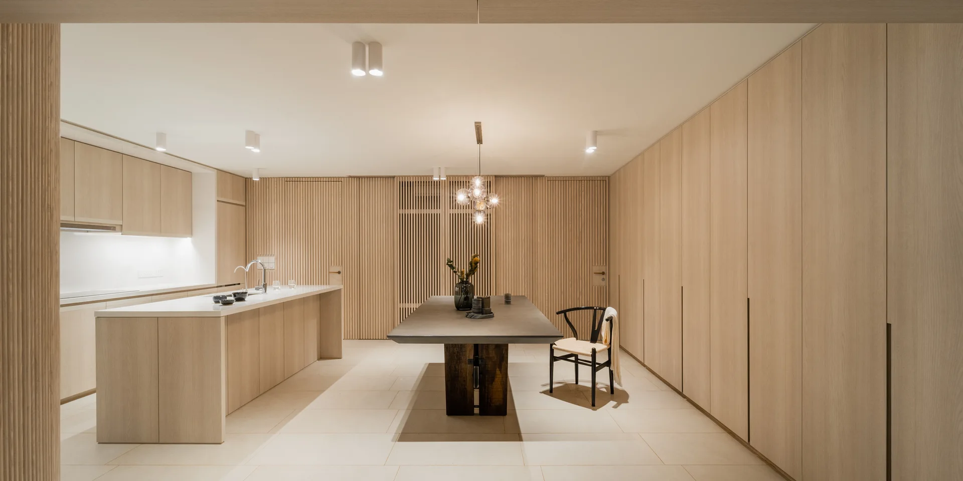 A minimalist kitchen and dining space featuring light wood slatted wall panels, a long dining table, a kitchen island, and a globe pendant light.