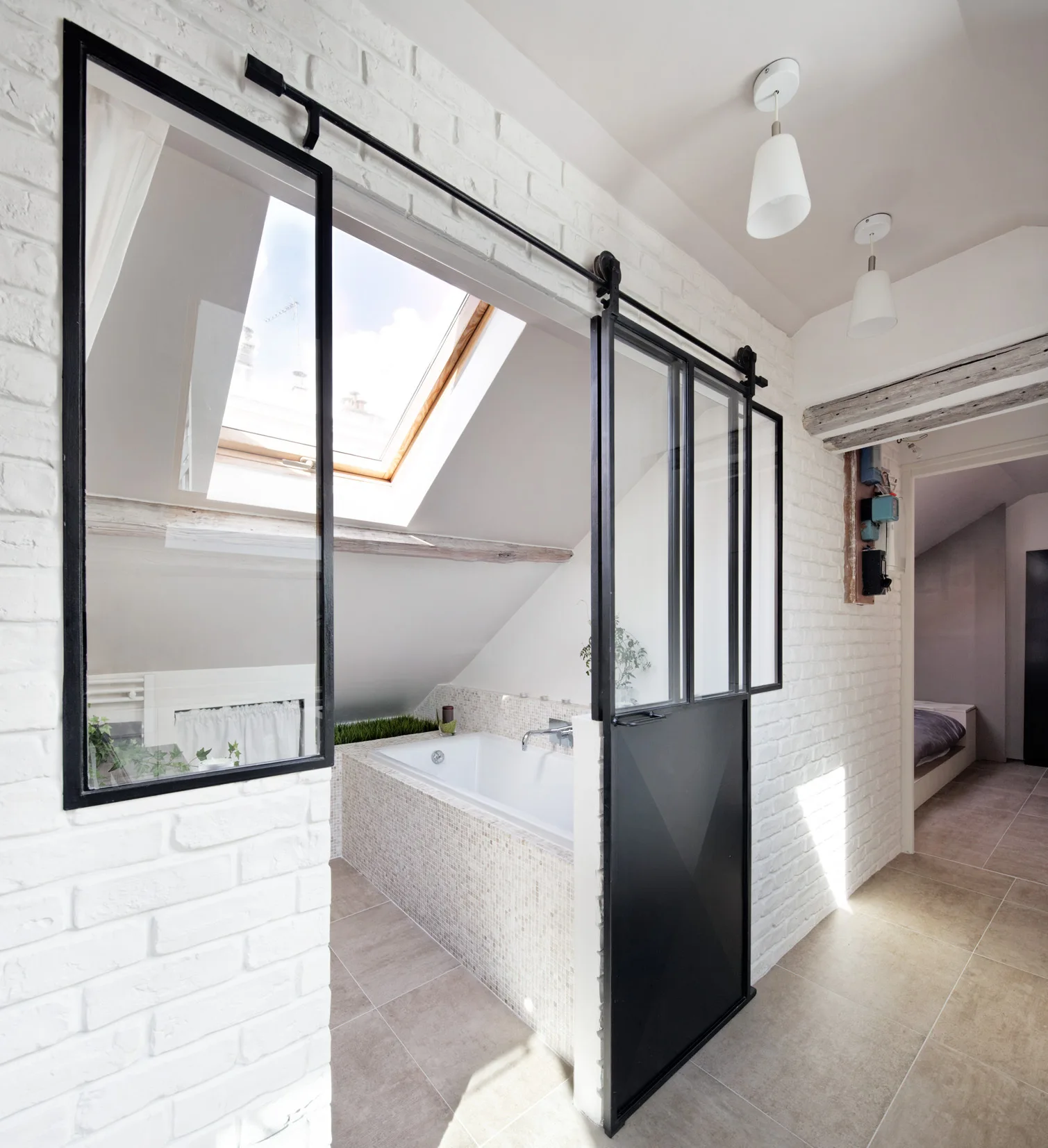 An attic bathroom features a built-in tub under a skylight and a black metal glass sliding door set against painted white brick.