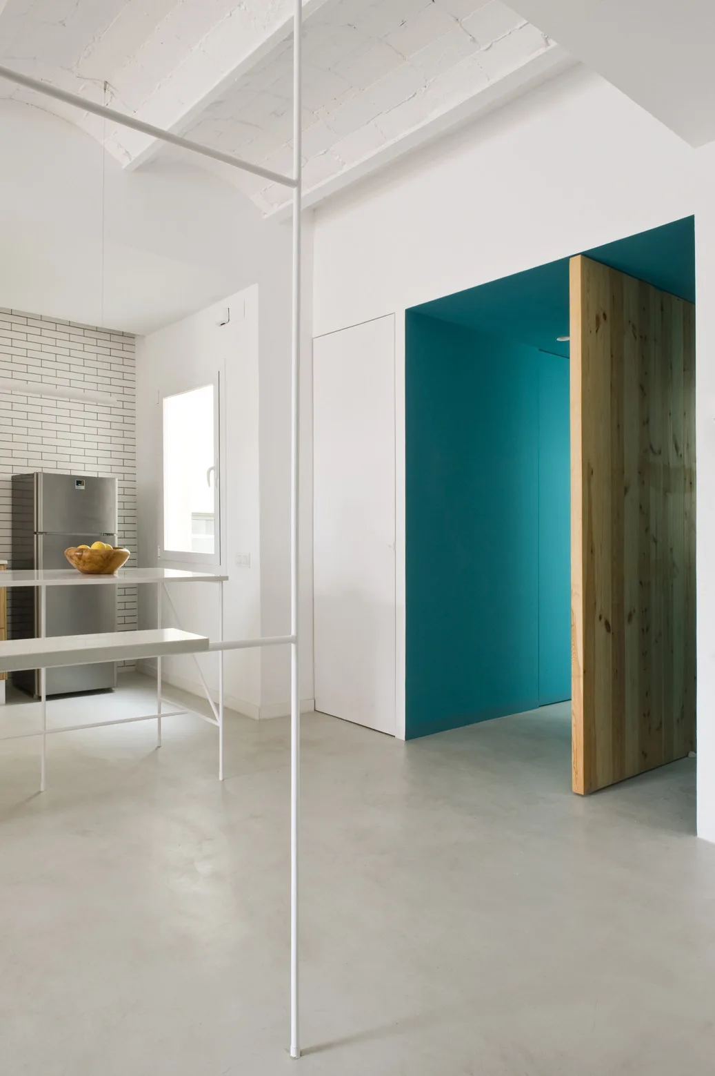 A minimalist white kitchen features a metal island frame, a stainless steel refrigerator, and a recessed blue passageway with a tall wood door.