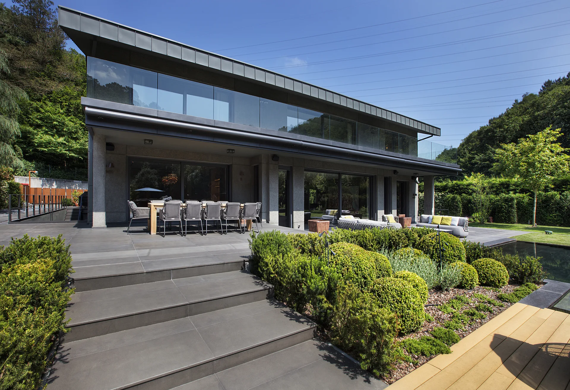 A modern two-story home features a large grey stone tile patio with outdoor dining and lounge furniture next to manicured shrub garden beds.