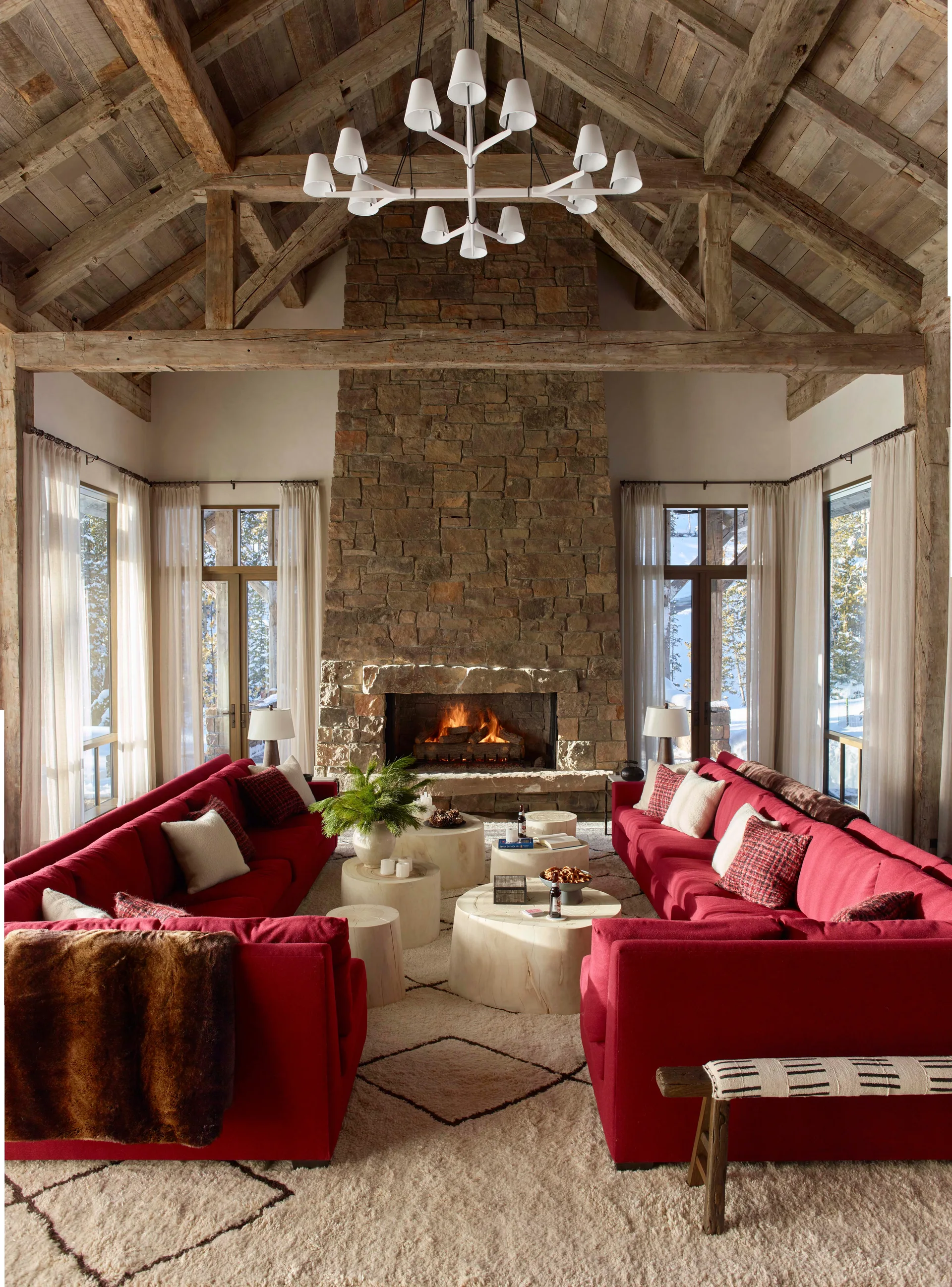 A rustic living room with a large stone fireplace, exposed timber vaulted ceilings, and two red sofas facing each other on a plush rug.