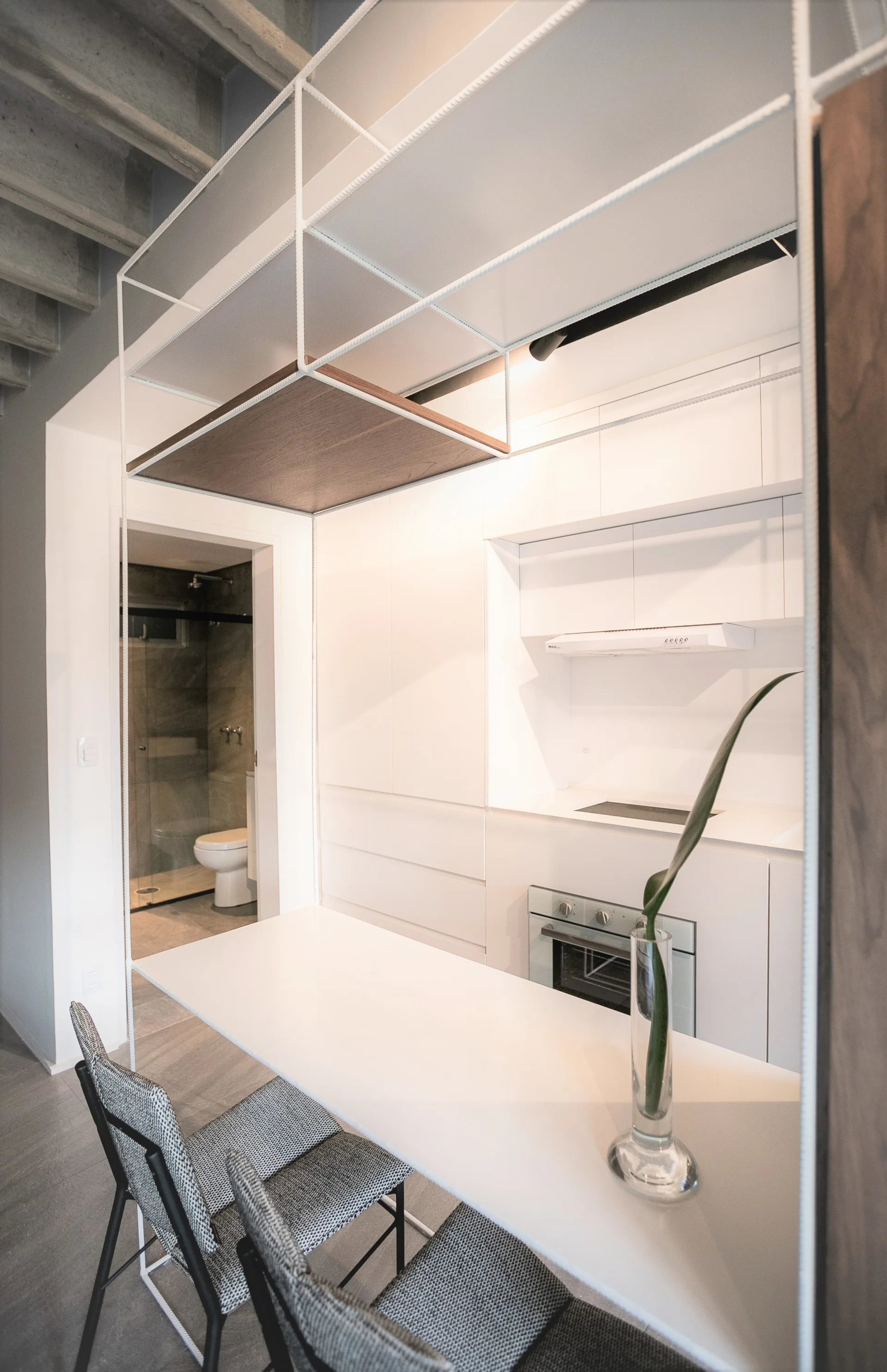 A compact white kitchenette features a white dining peninsula with grey chairs under hanging white metal grid shelves against a concrete ceiling.