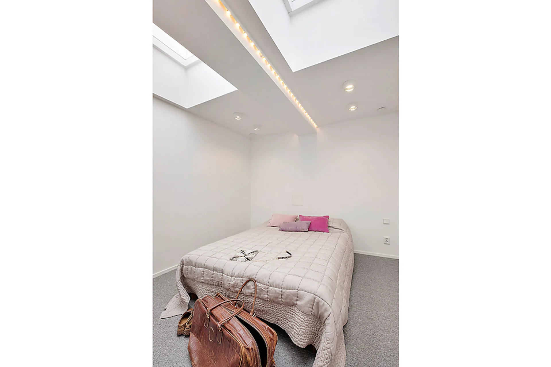 A minimalist bedroom with white walls, a bed featuring a quilted coverlet and pink pillows, grey carpet, and overhead skylights.