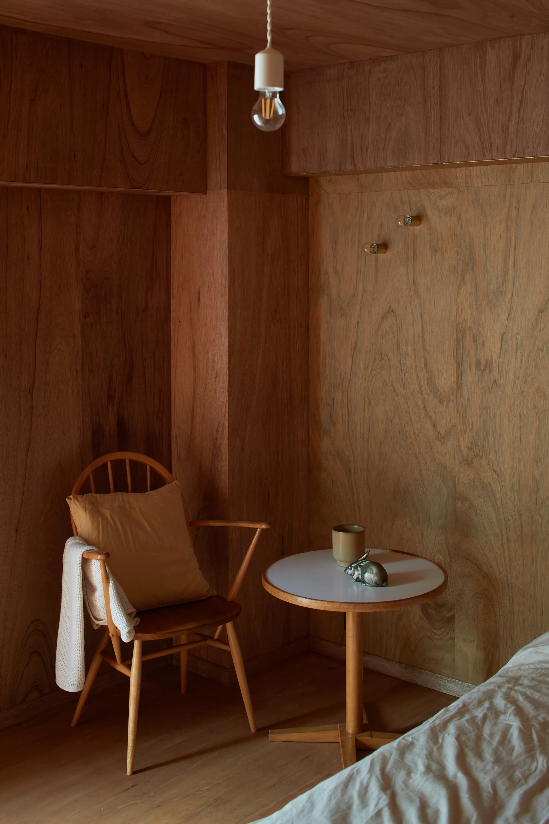 A minimalist bedroom nook features warm plywood paneled walls, a wooden spindle chair with a tan cushion, and a small round white-topped pedestal table.