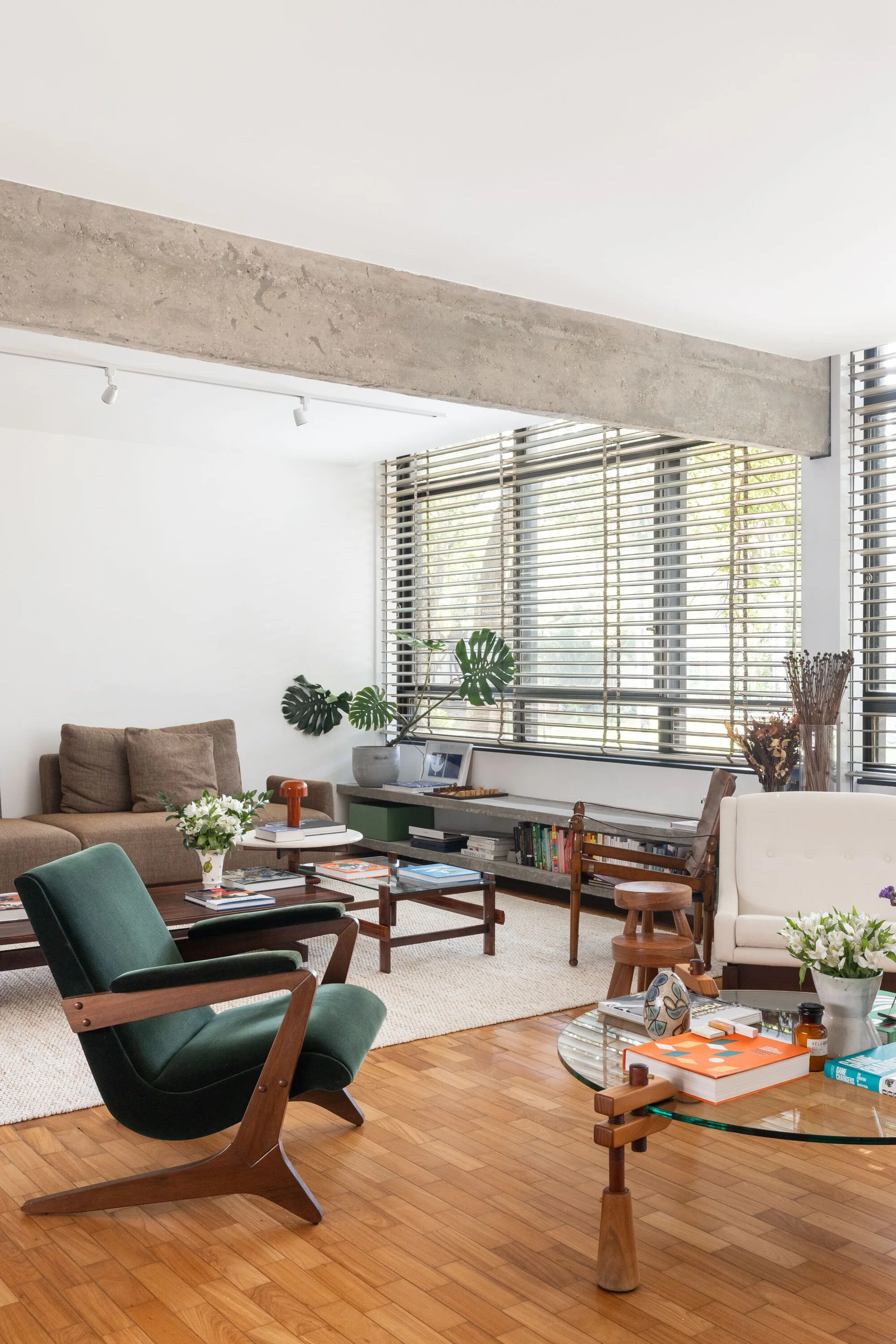 A mid-century living room features a green velvet armchair, wood parquet floors, an exposed concrete ceiling beam, and large window blinds.