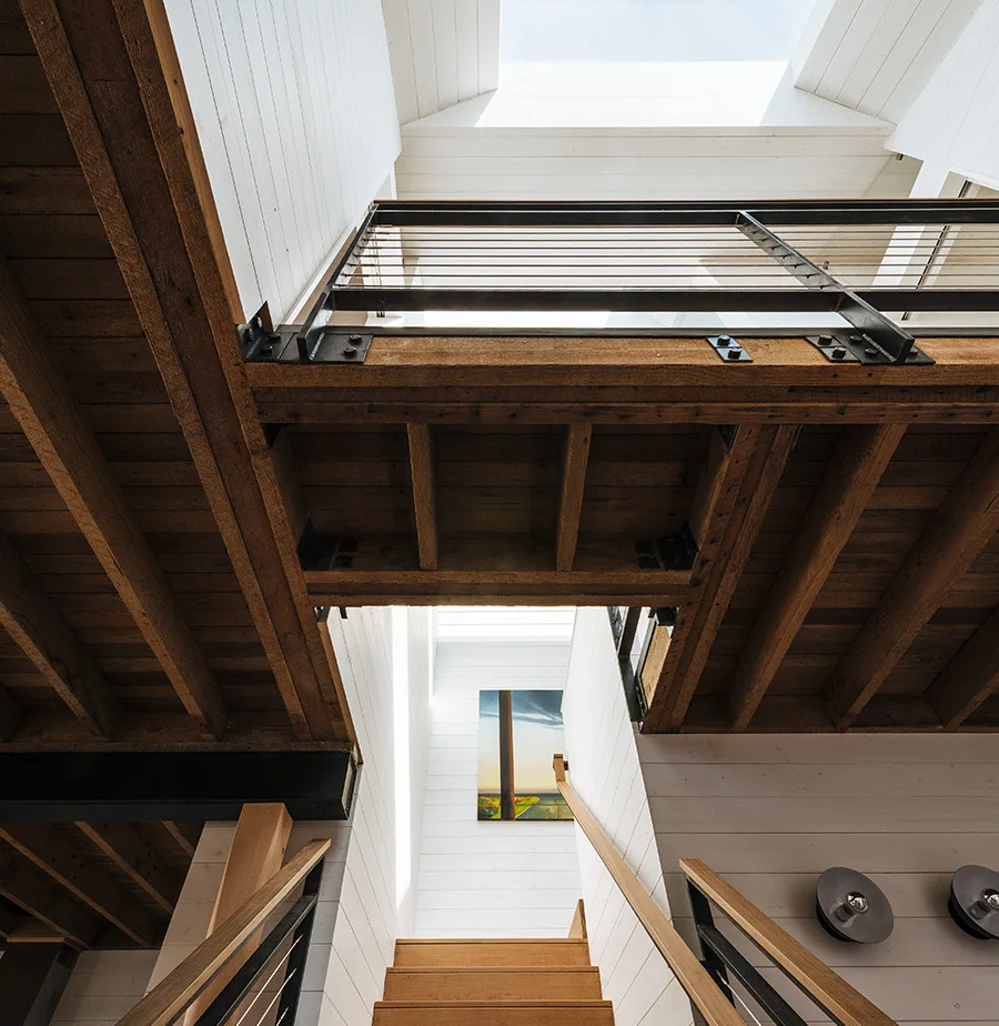 Looking up a wooden staircase with metal cable railings, white shiplap walls, and exposed ceiling beams illuminated by a bright skylight.