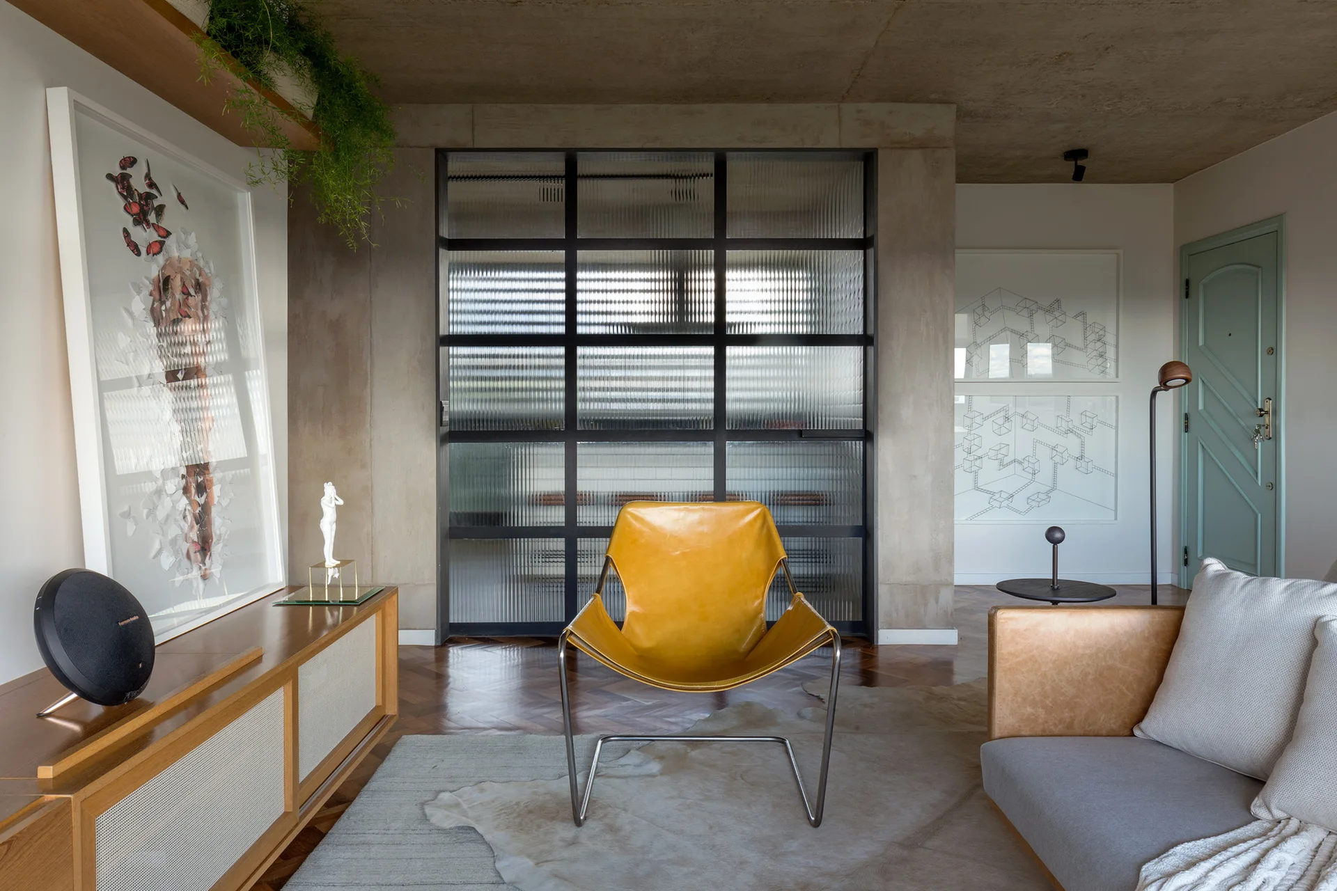 A modern living room features a yellow leather armchair in front of a fluted glass partition, concrete ceiling, and herringbone wood floor.
