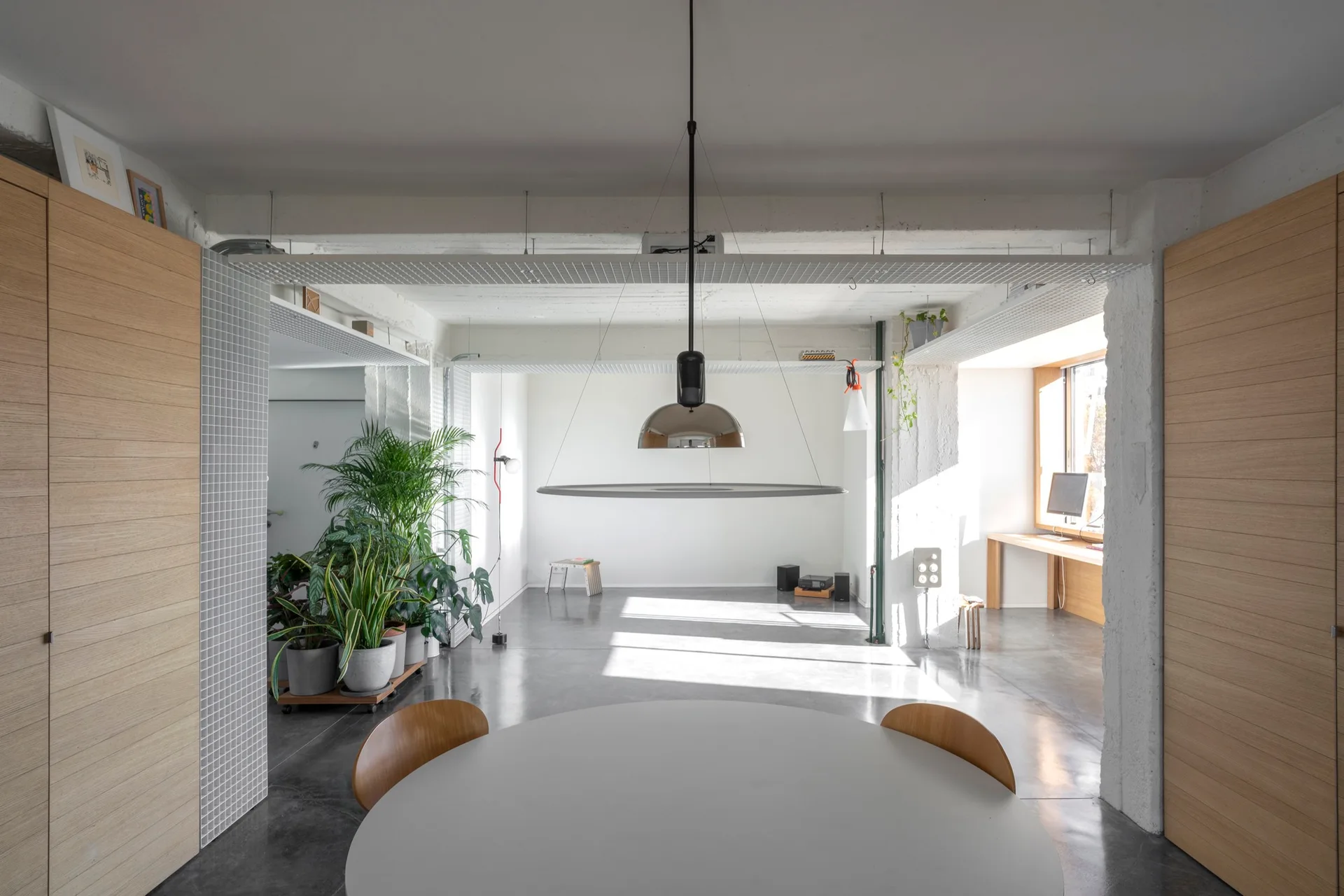 A minimalist open-plan dining space features a round white table, concrete floors, wood-paneled walls, and overhead industrial metal grids.