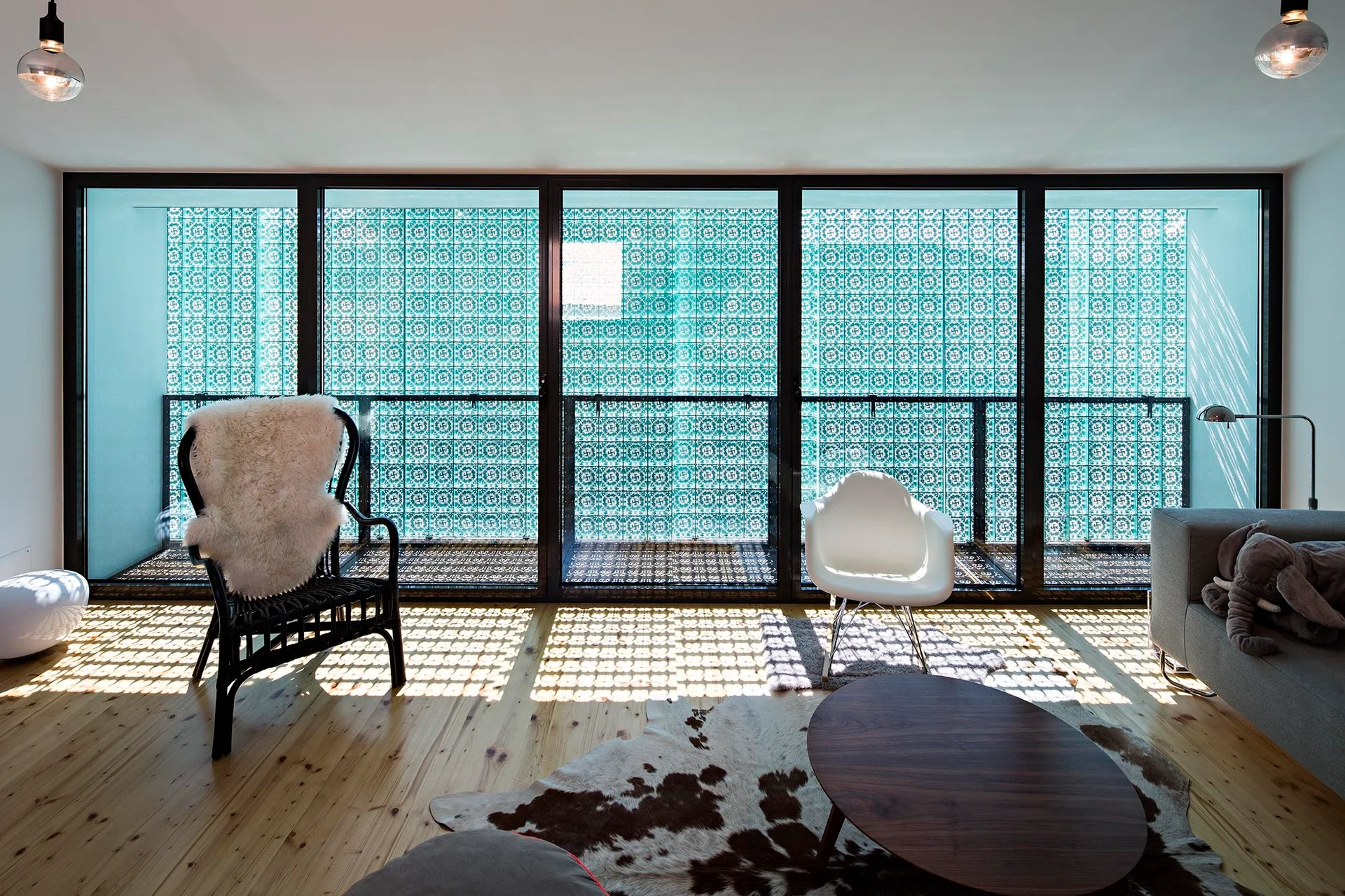 An airy living room with large glass doors showing teal geometric screens that cast patterned shadows across light wood floors and eclectic chairs.