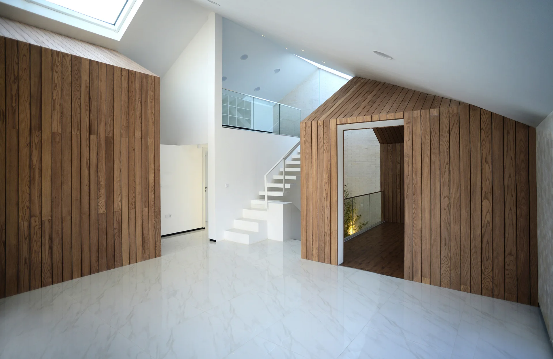 A minimalist double-height hallway features house-shaped wooden structures, polished marble floors, and a white floating staircase under a skylight.