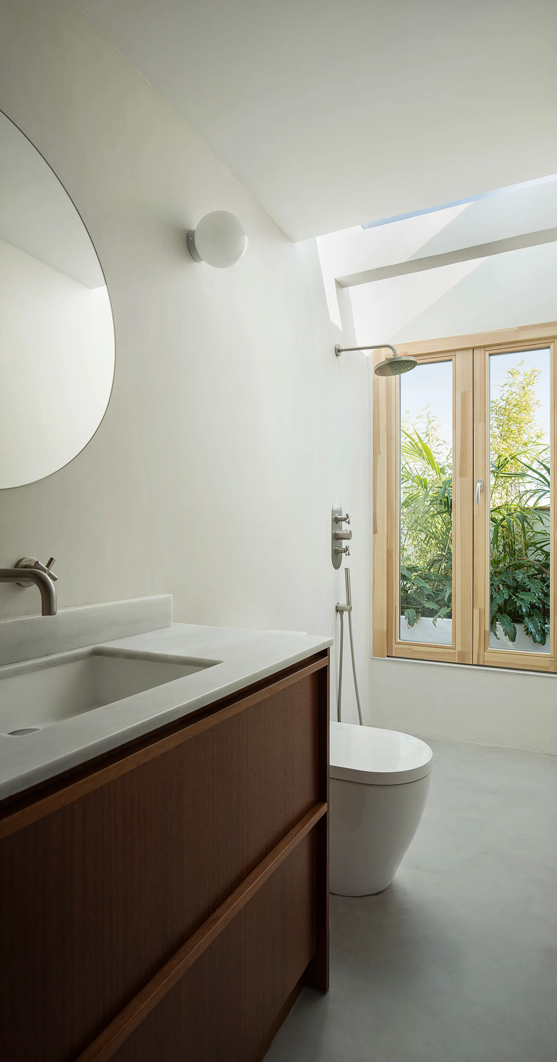 A minimalist bathroom featuring a dark wood vanity, round mirror, white toilet, and a wood-framed window looking out to green foliage.