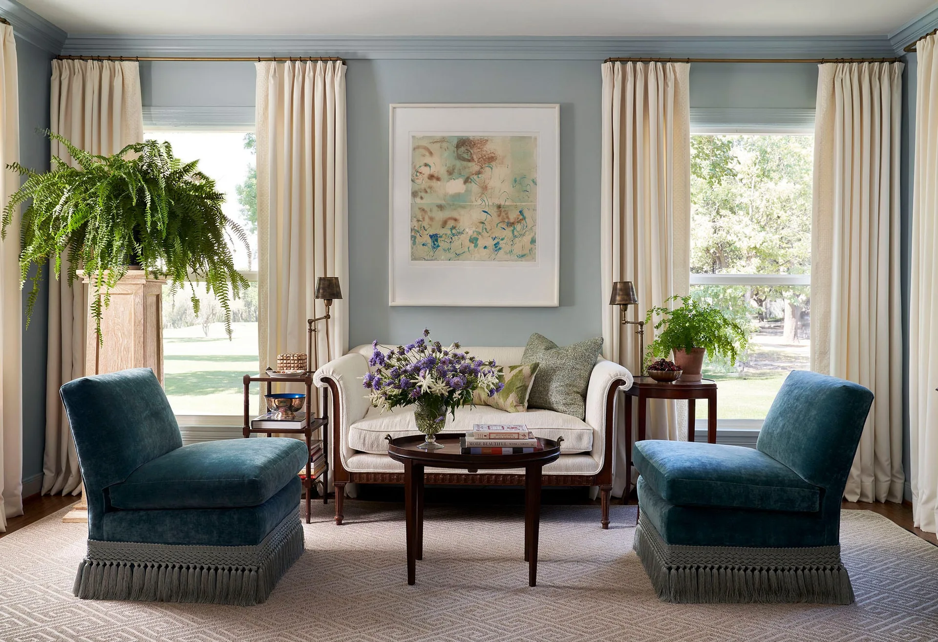 A symmetrical blue living room features a central cream sofa flanked by windows, a dark wood coffee table, and two teal fringed armchairs.