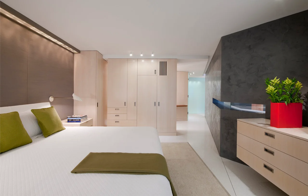 A modern bedroom featuring built-in light wood cabinetry, a white bed with green accent pillows, and a dark textured wall.