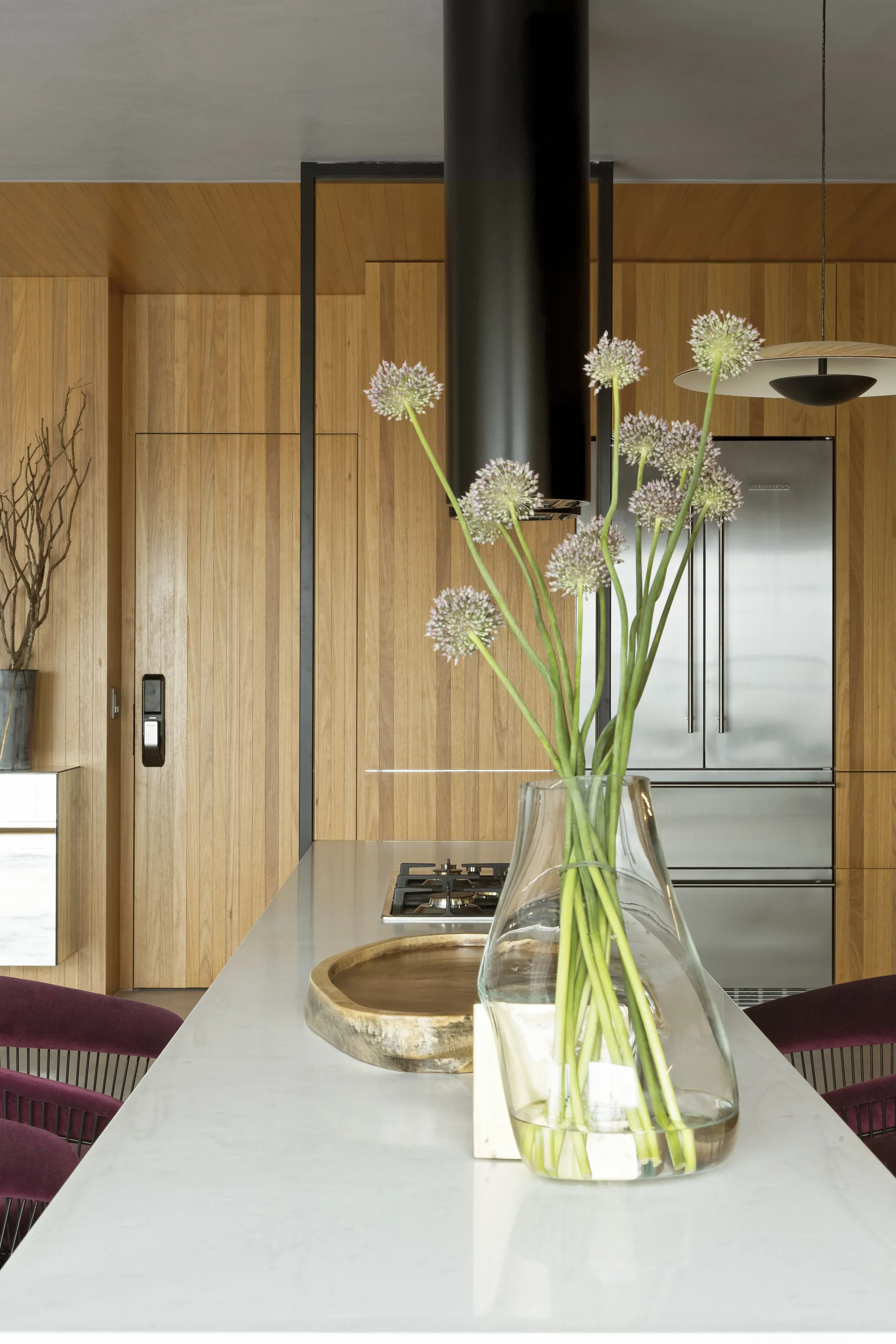 A modern kitchen featuring a stone-topped island with purple chairs, set against vertical wood-paneled walls and a stainless steel refrigerator.