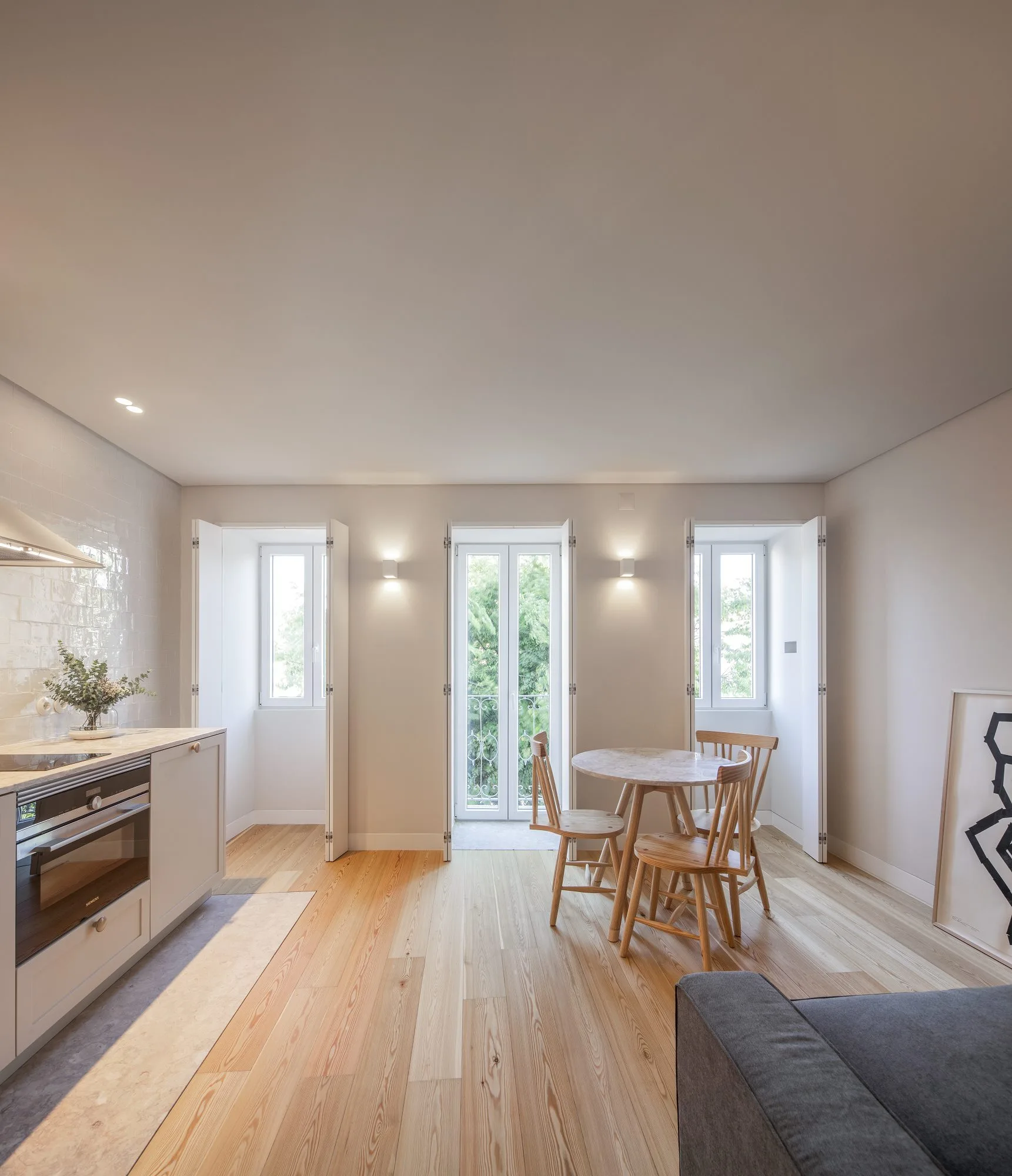 A bright dining and kitchen space features a round wooden table, beige cabinetry, and three deep window bays with folding white shutters.