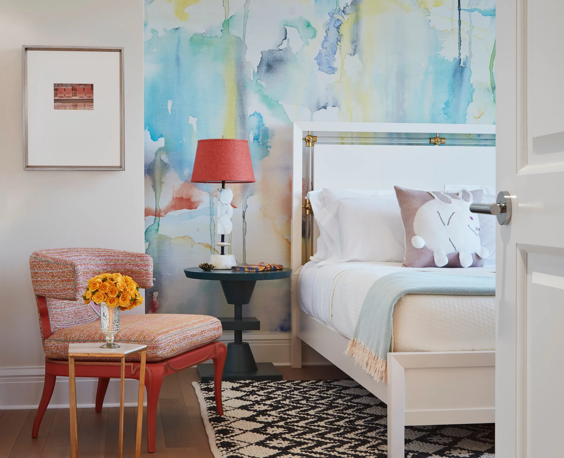 A contemporary bedroom features a watercolor wall mural behind a white bed, with a coral armchair and a black pedestal nightstand nearby.