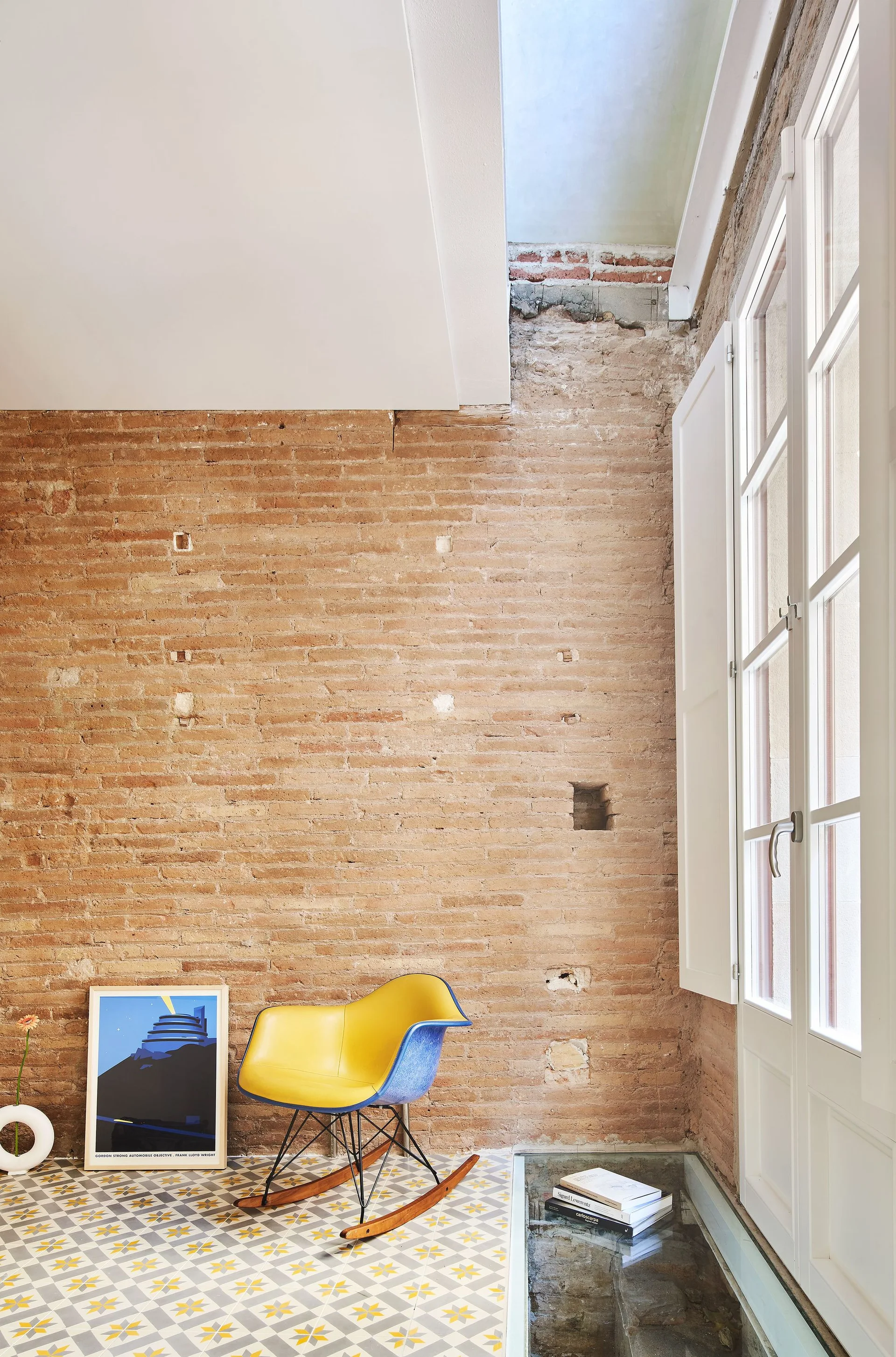 A minimalist room corner features an exposed brick wall, a yellow molded rocking chair on a patterned tile floor, and a tall white window.