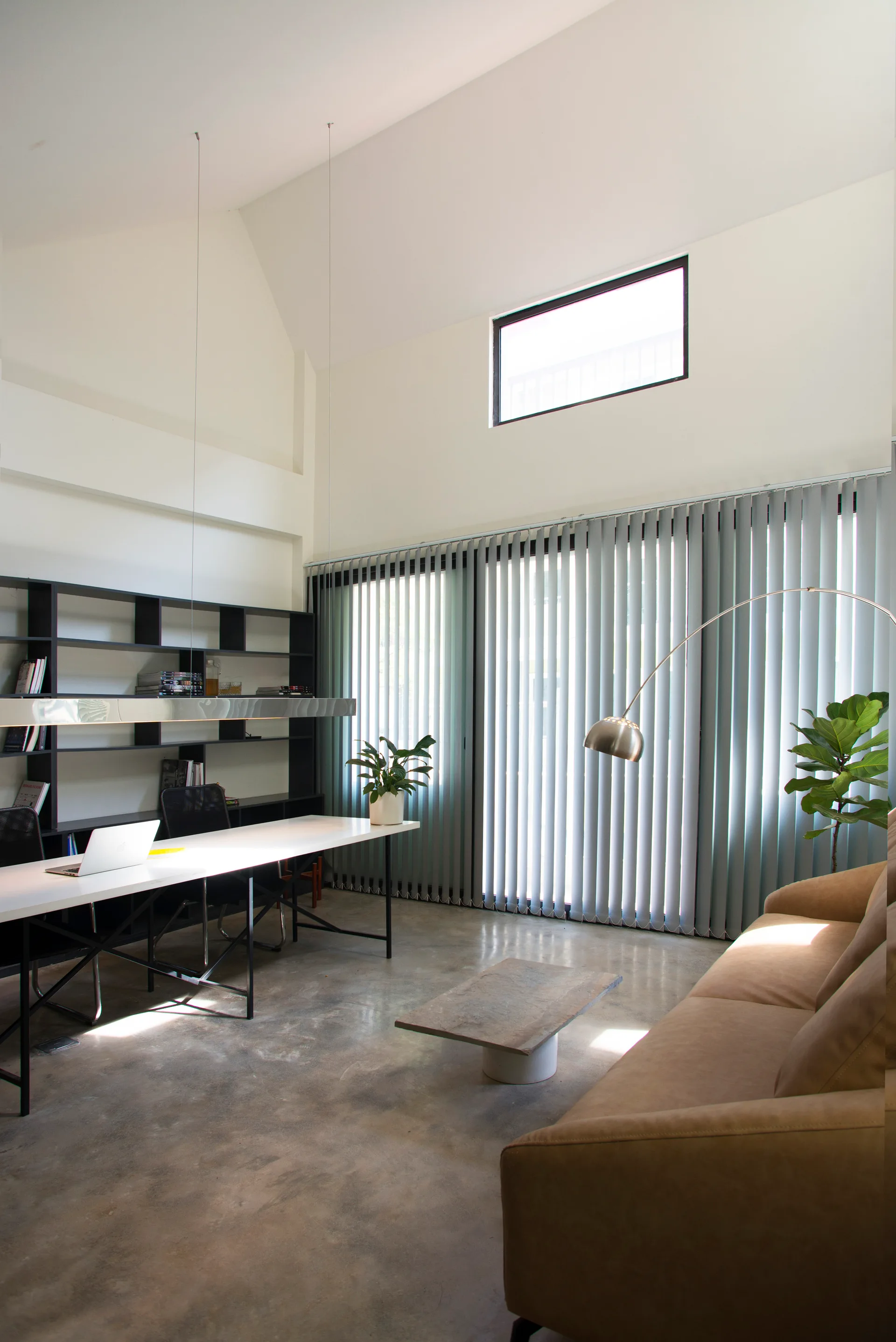 A modern home office featuring a high vaulted ceiling, polished concrete floor, a tan sofa, and a long white desk against black shelving.