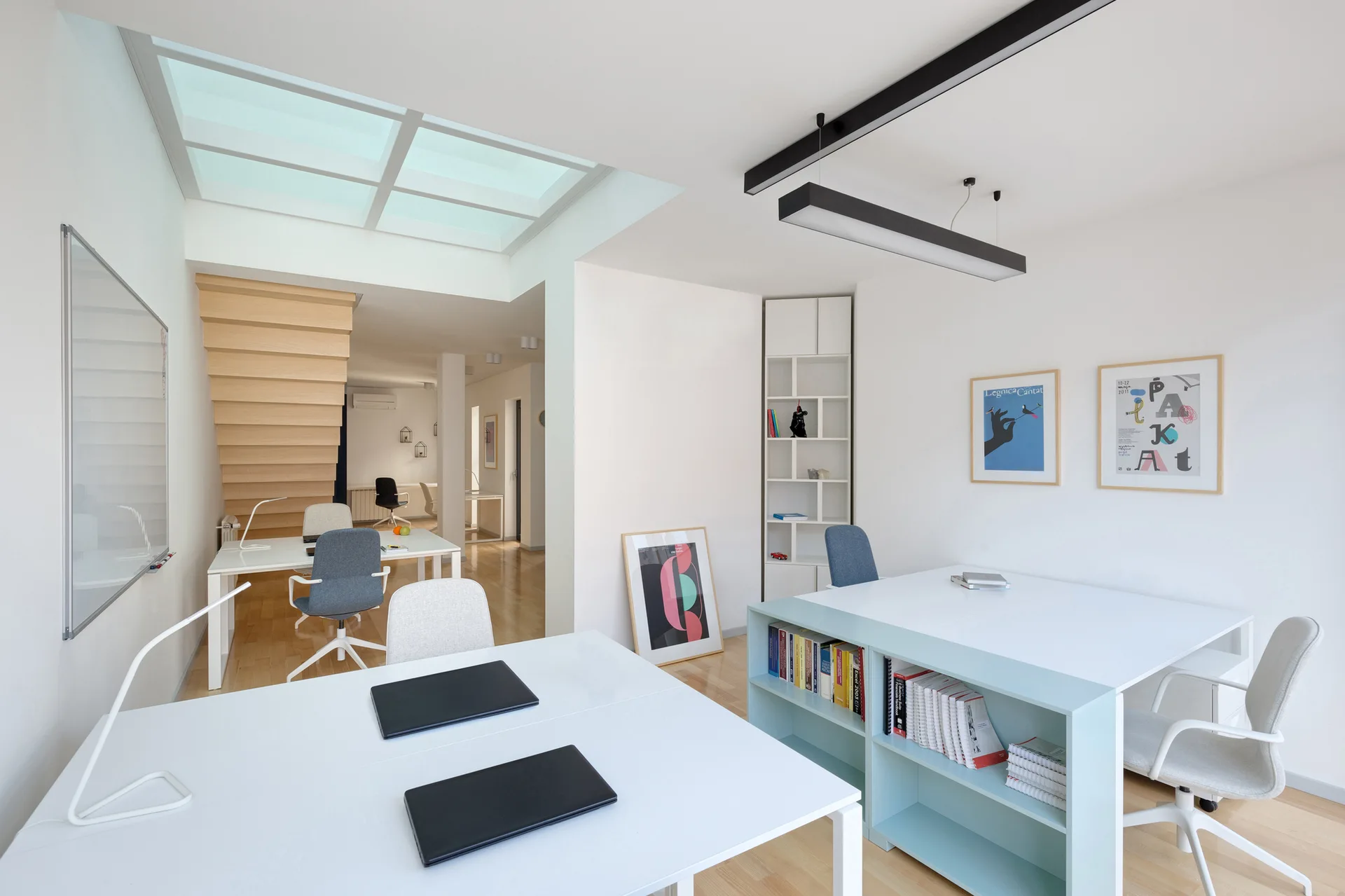 A modern white workspace featuring multiple desks with grey office chairs, a wooden staircase in the background, and a bright overhead skylight.