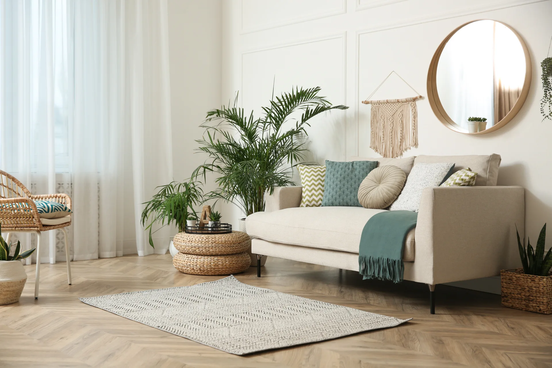 A beige sofa with green and teal pillows sits on herringbone wood floors next to a round mirror and potted houseplants.