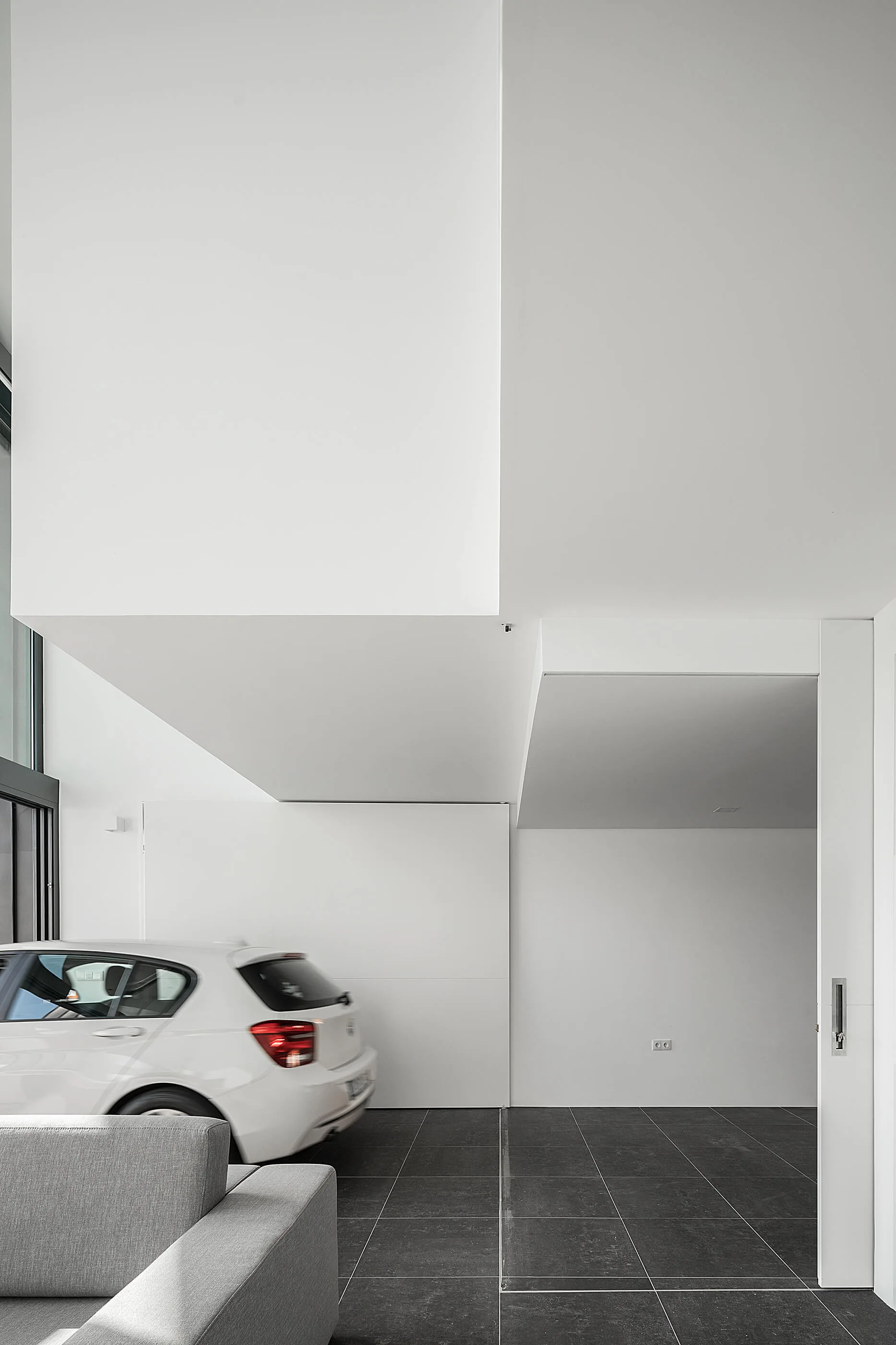 A grey sofa sits in a minimalist white living room with dark gray tile flooring next to a white car parked indoors.
