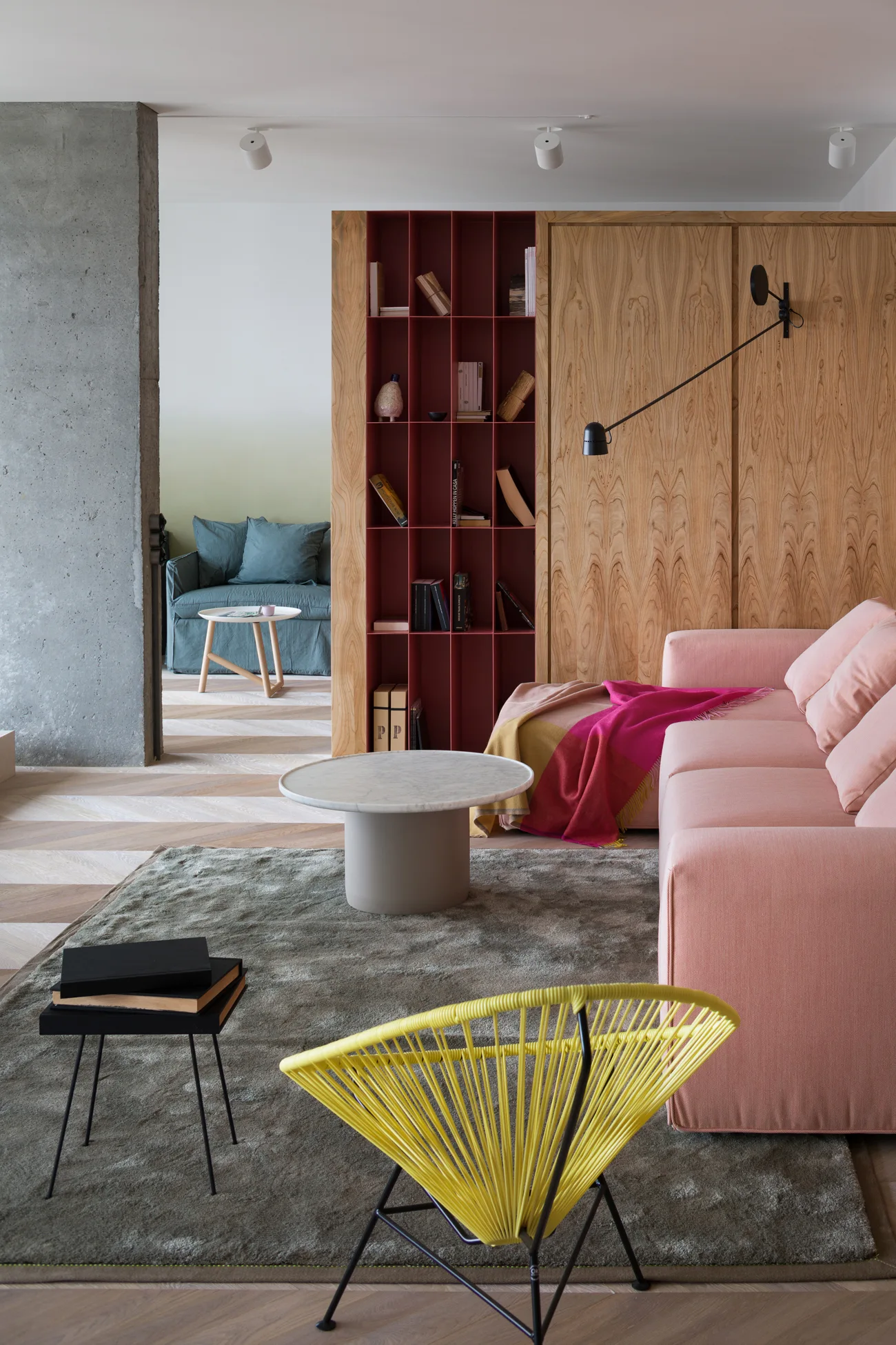 A contemporary living room featuring a pink sofa, a yellow wire accent chair, a grey area rug, and a custom wood room divider with red shelving.