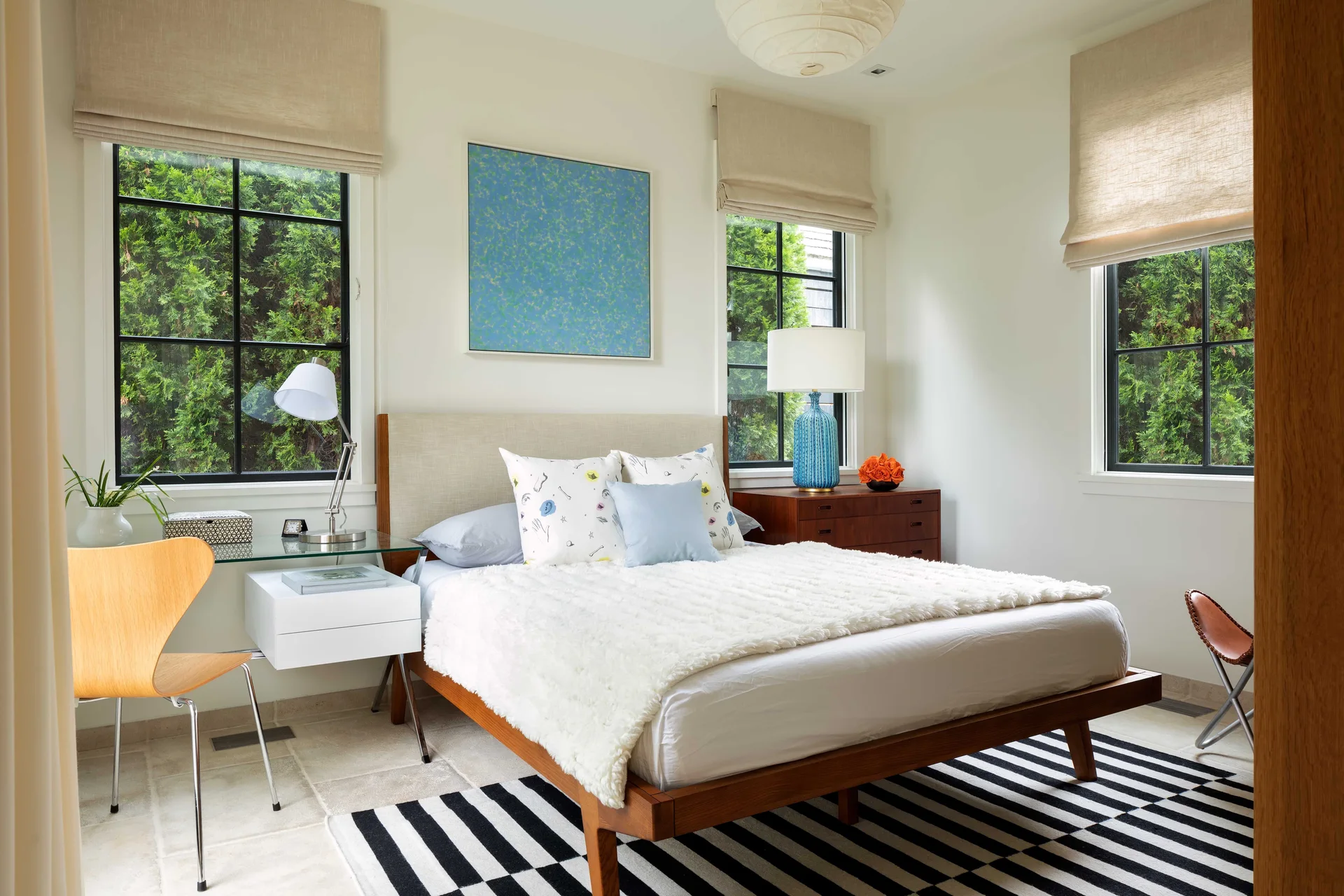 A bright bedroom features a wooden bed, a black and white striped rug, a glass-topped desk, and three windows with linen roman shades.