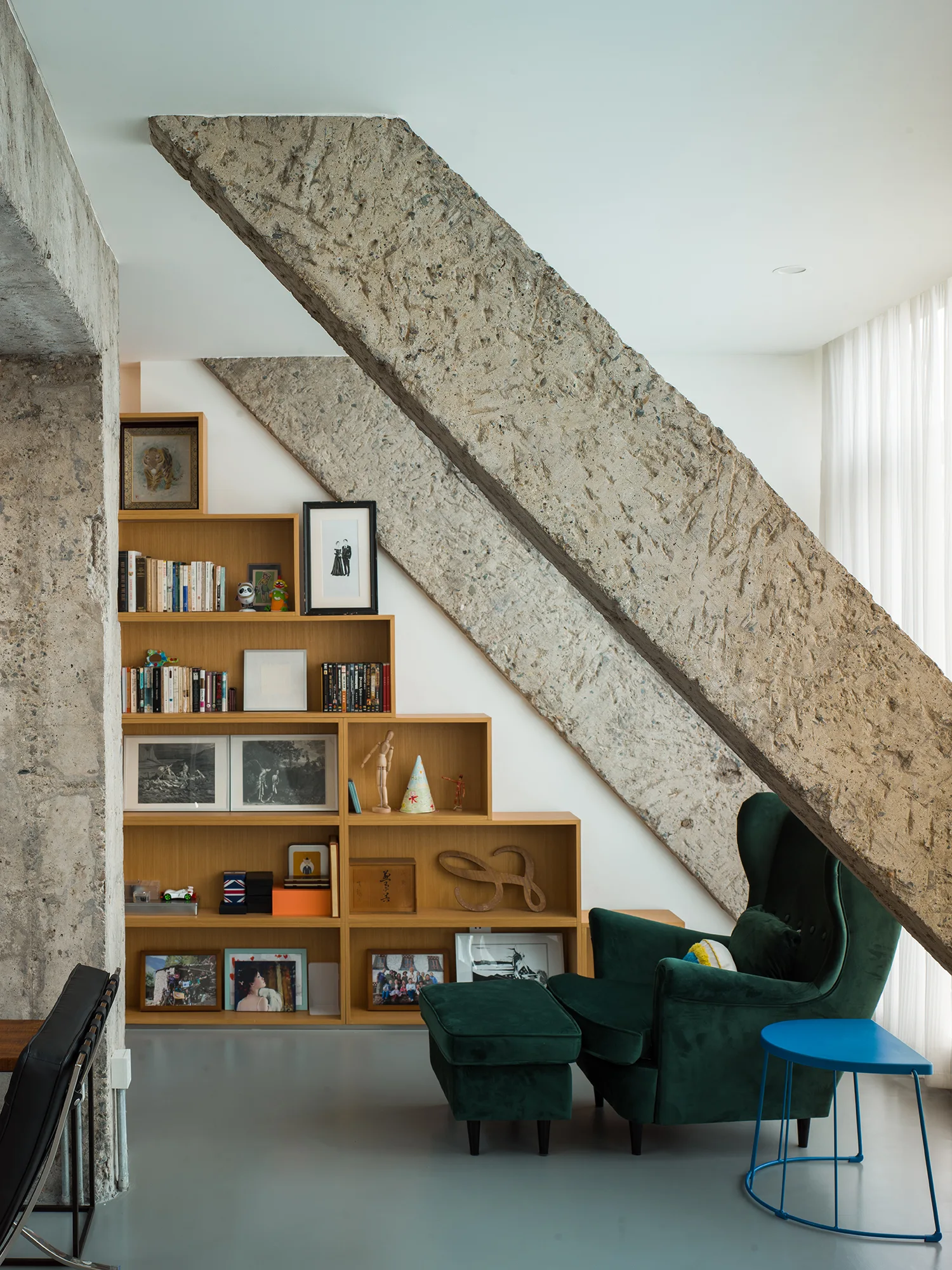A green velvet wingback chair and ottoman sit next to a stepped wooden bookshelf nestled beneath heavy, diagonal raw concrete structural beams.