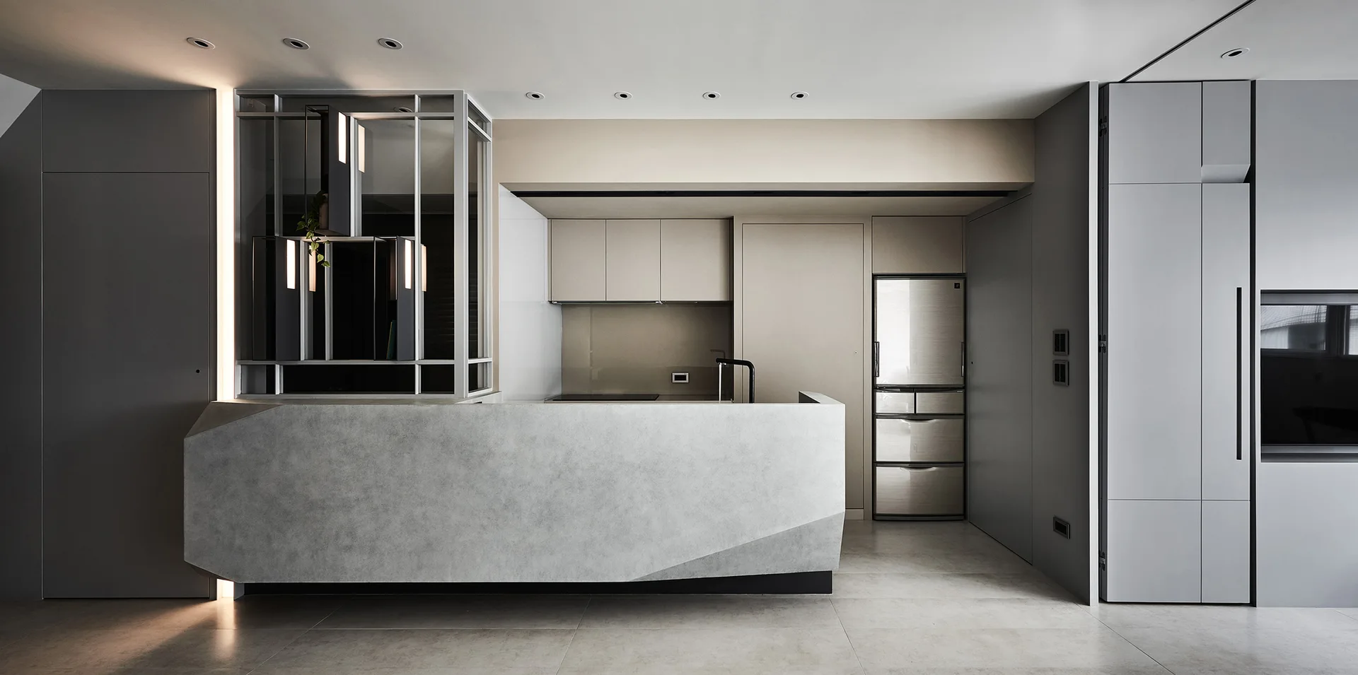 A minimalist kitchen features a sculptural grey concrete island, light beige cabinets, a stainless steel refrigerator, and a black faucet.