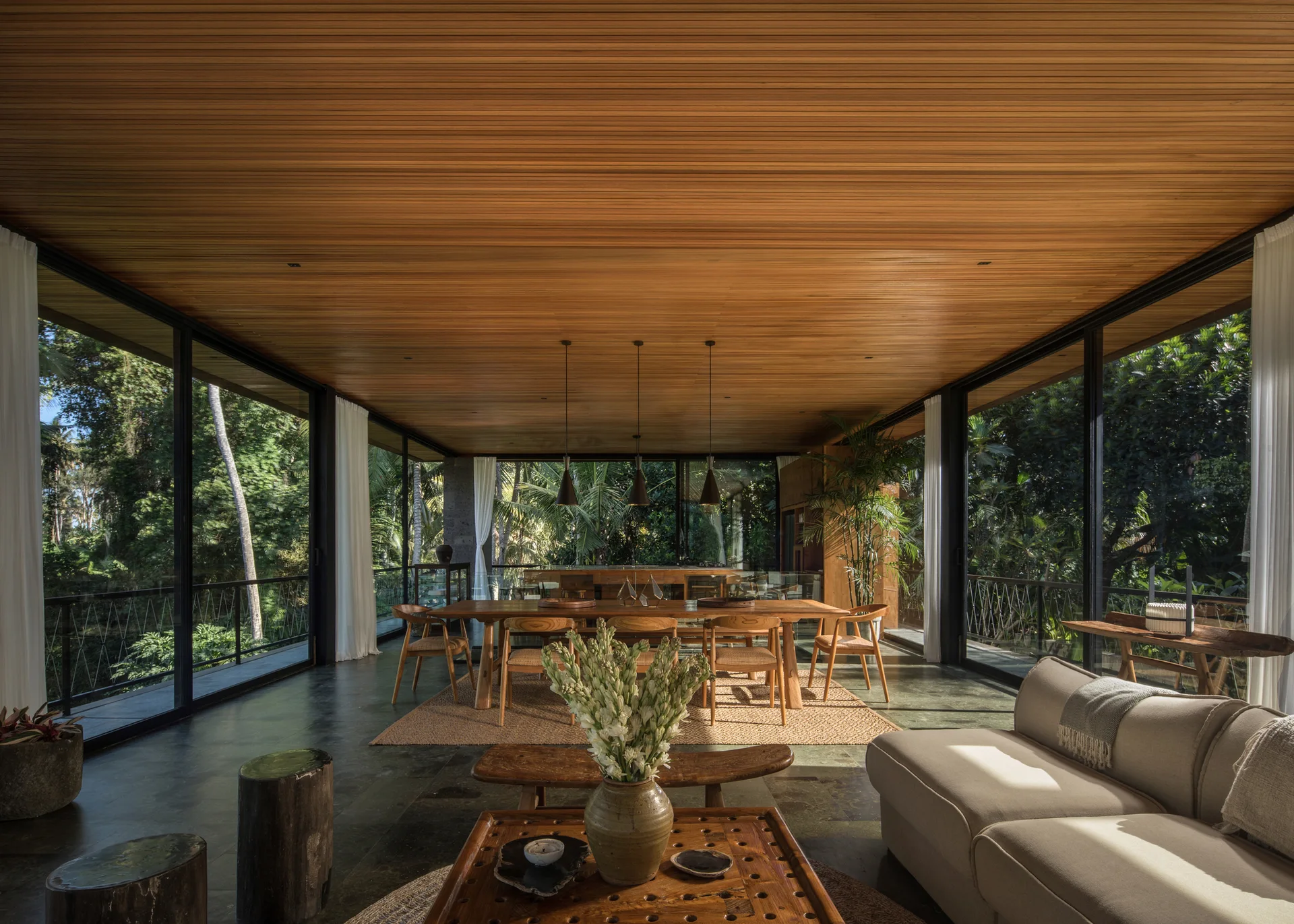 A modern open-concept dining room features a wood-slat ceiling, dark stone flooring, and floor-to-ceiling windows showing a tropical forest.