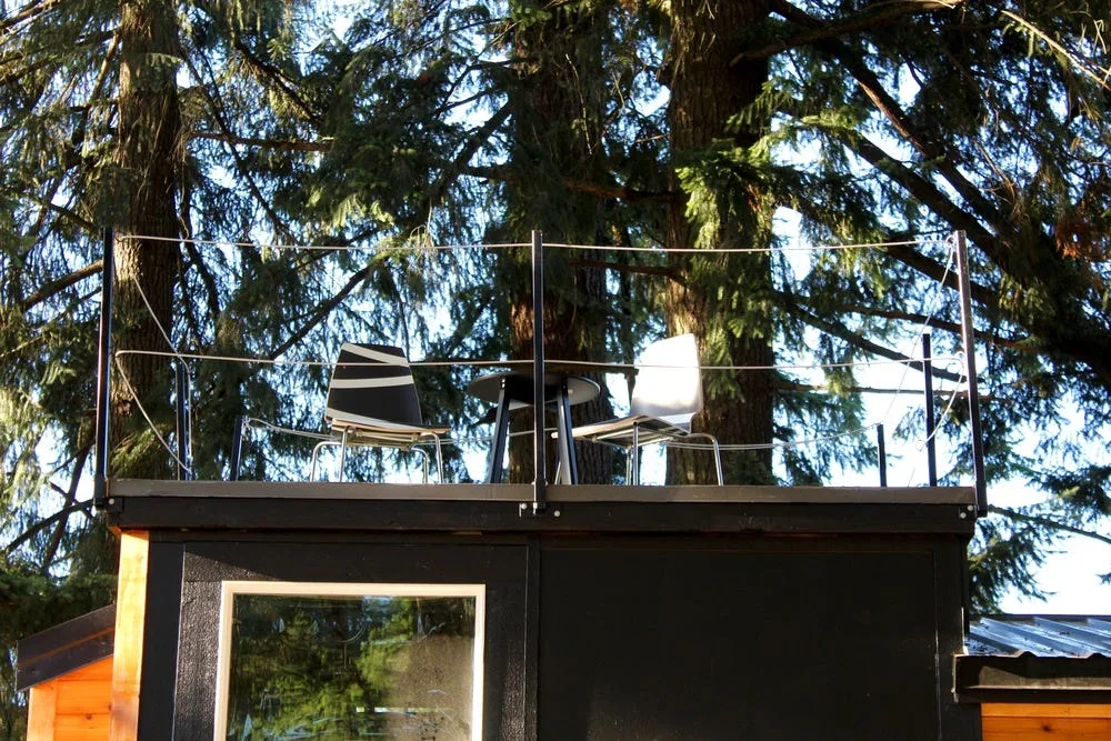 An elevated black deck features a modern cable railing, a small round table, and two chairs set against a dense forest of evergreen trees.