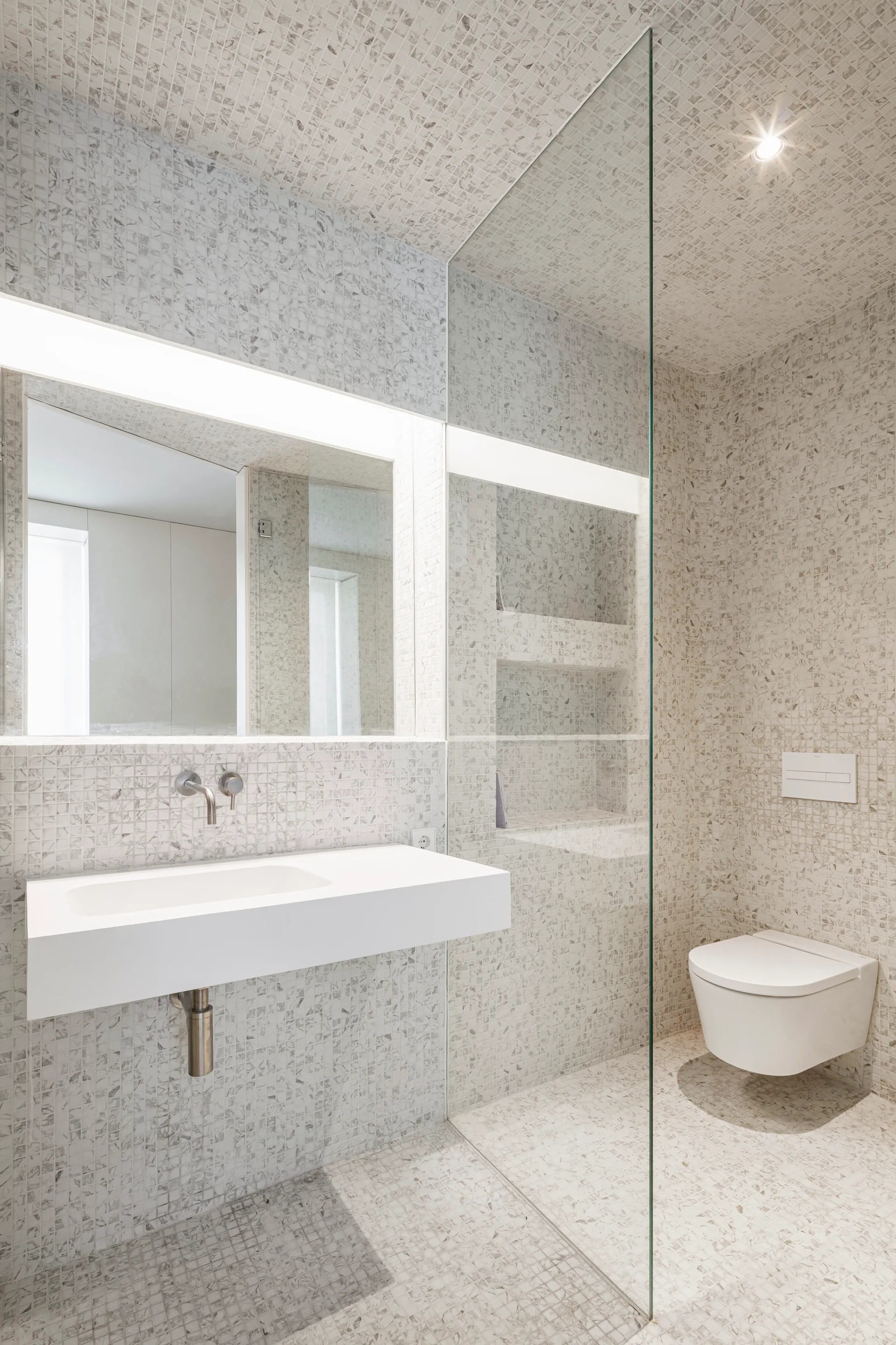 A minimalist bathroom features gray and white mosaic tiling on the walls, floor, and ceiling, a floating white sink, and a glass shower partition.