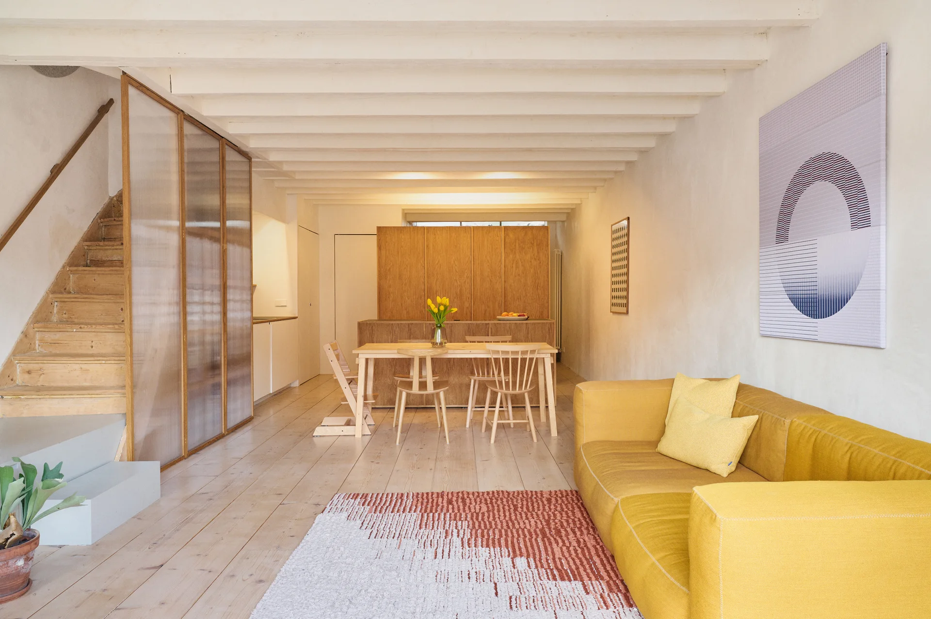 A minimalist open-plan living and dining room features a bright yellow sofa, a light wood dining table, and a translucent screen next to a wooden staircase.