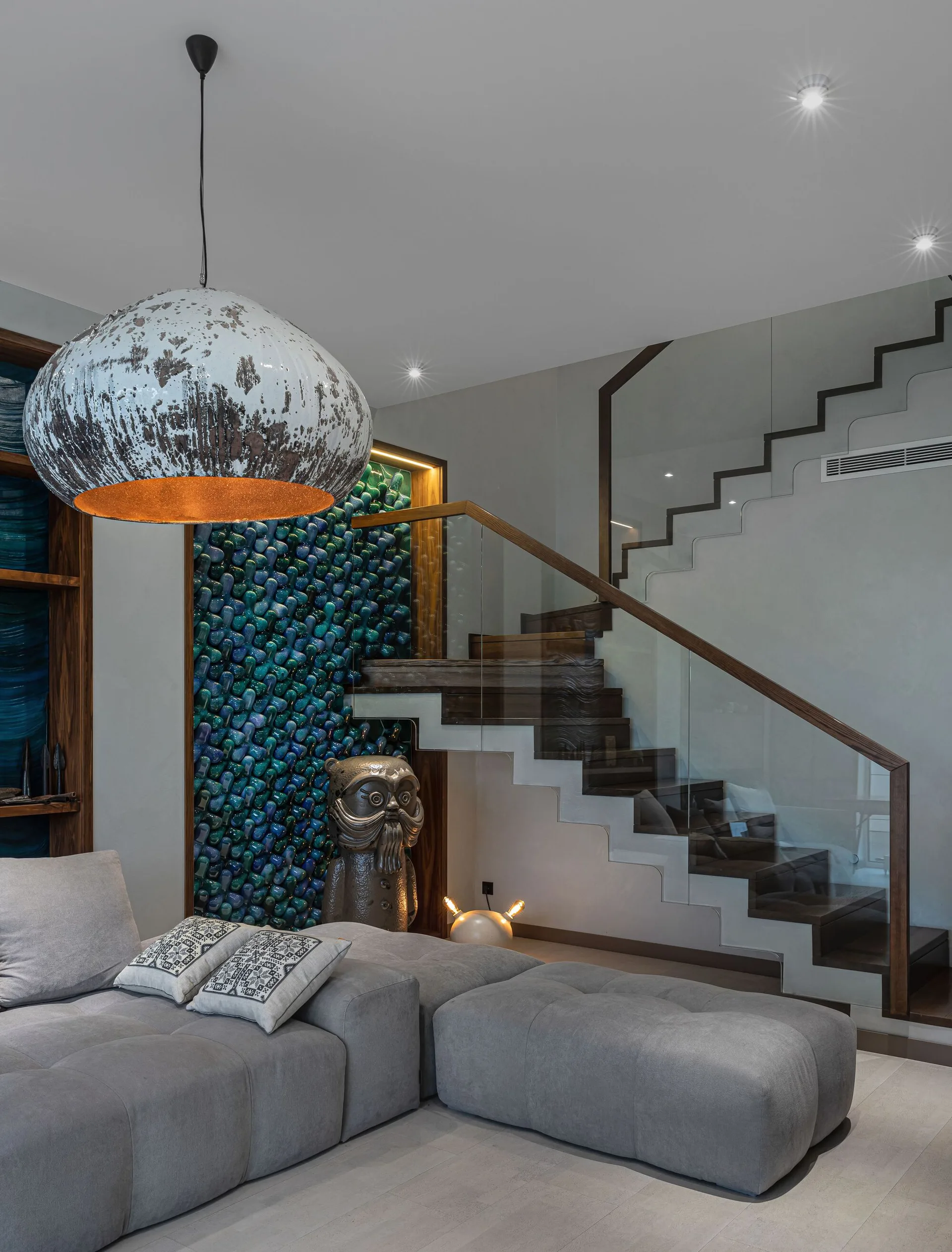 A modern grey sectional sofa is positioned near a floating wooden staircase with glass railings and a textured blue accent wall.