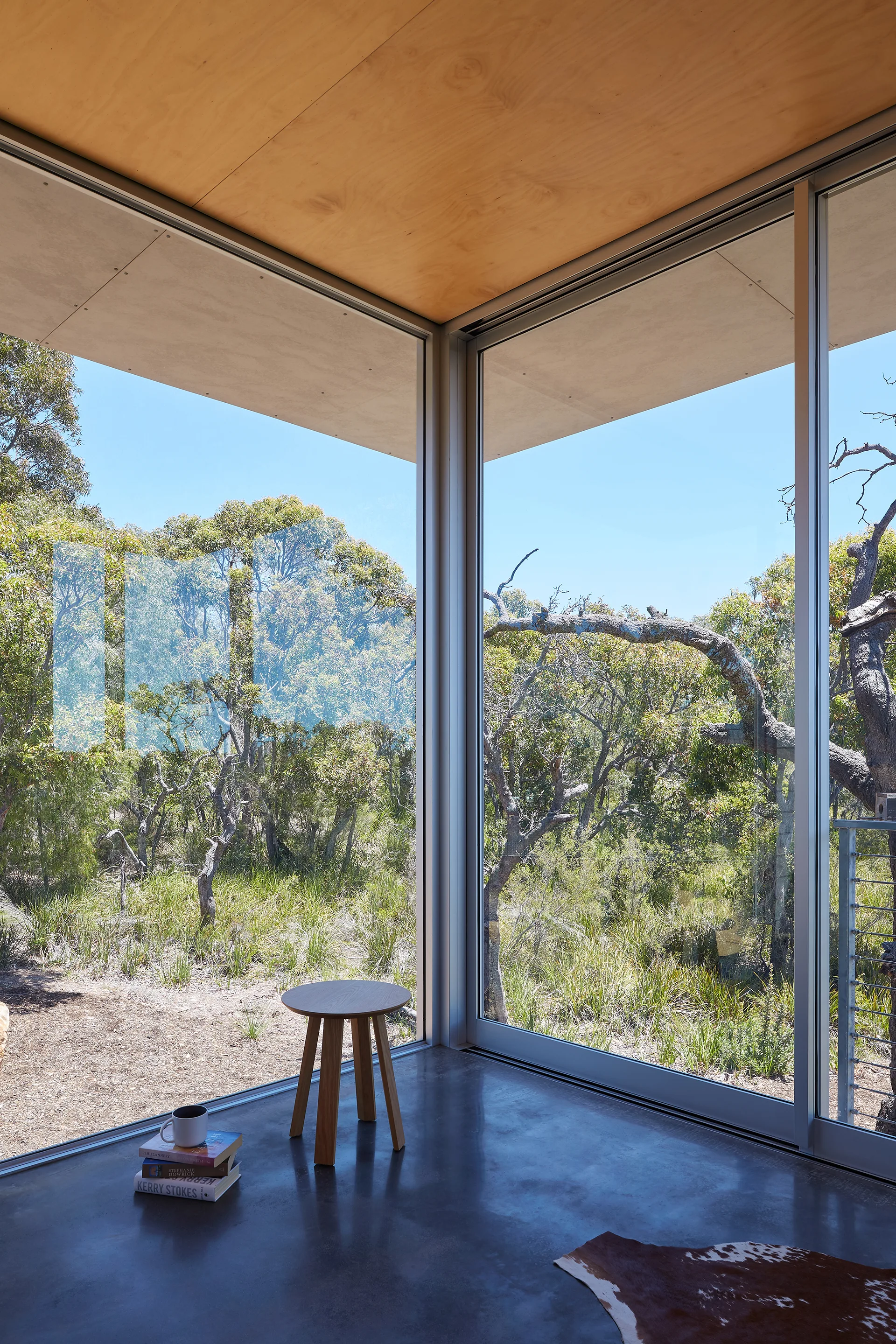 A minimalist room corner features floor-to-ceiling glass walls overlooking a forest, with a wood stool and book stack on a polished concrete floor.