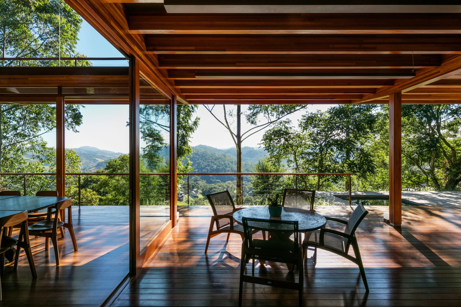 A covered wooden terrace features dining sets under an exposed beam ceiling next to large sliding glass doors looking out at green hills.