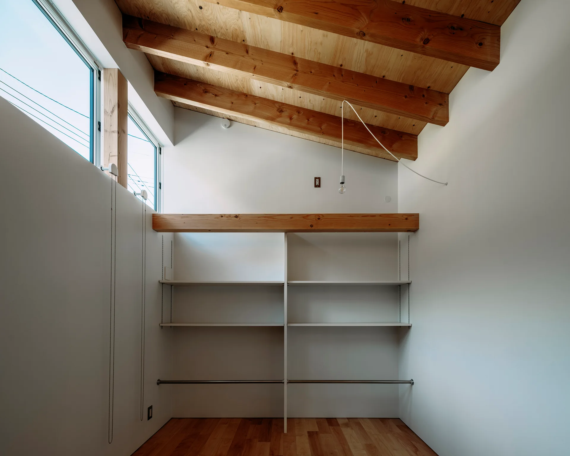 A minimalist closet space featuring white built-in shelving, a metal clothes rod, light wood floors, and a sloped ceiling with exposed wood beams.